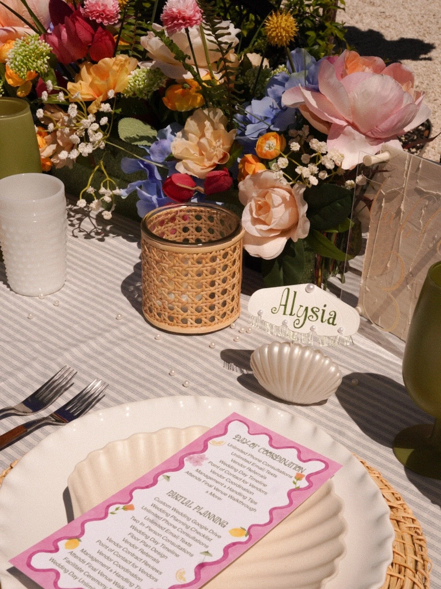 Table decorated with a bouquet of colorful flowers including roses, hydrangeas, and baby’s breath. There are plates, silverware, and a pink wedding program on the table. The table has a white and gray striped cloth, and there are small pearl-like beads scattered on it. A white name card with the name 'Alysia' is also visible.