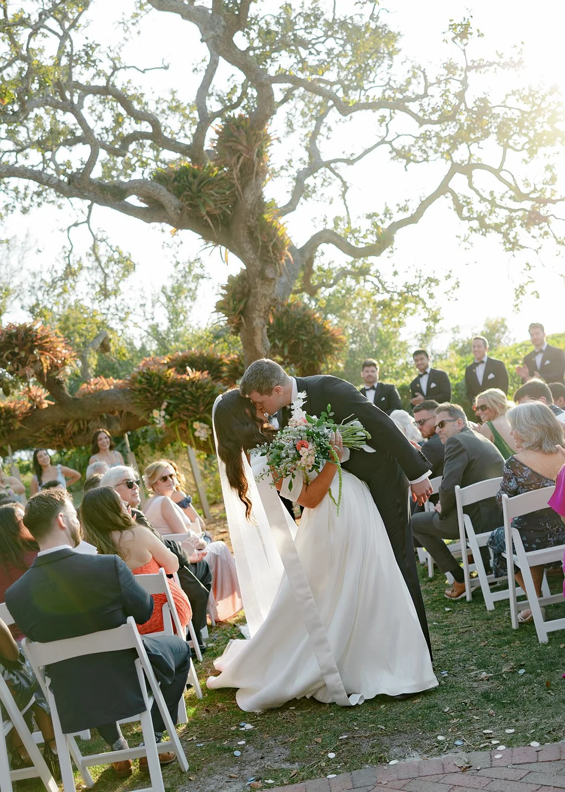 A bride and groom sharing a kiss during their outdoor wedding ceremony, surrounded by seated guests and a large tree in the background.