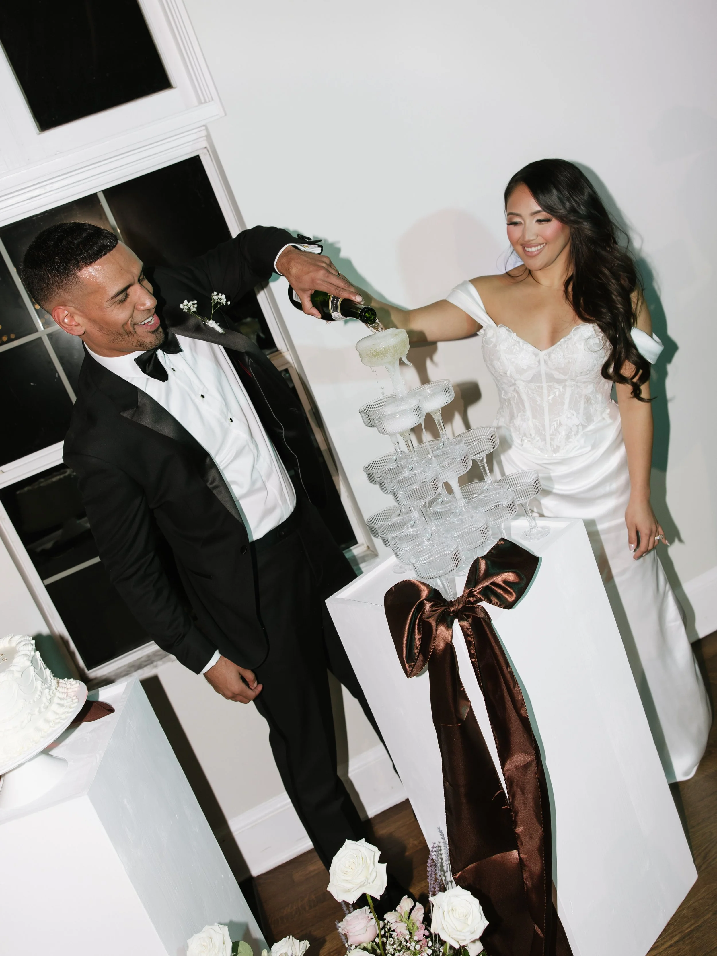 A bride and groom in wedding attire pour champagne into a tower of champagne glasses at their wedding celebration, with flowers and a cake nearby.