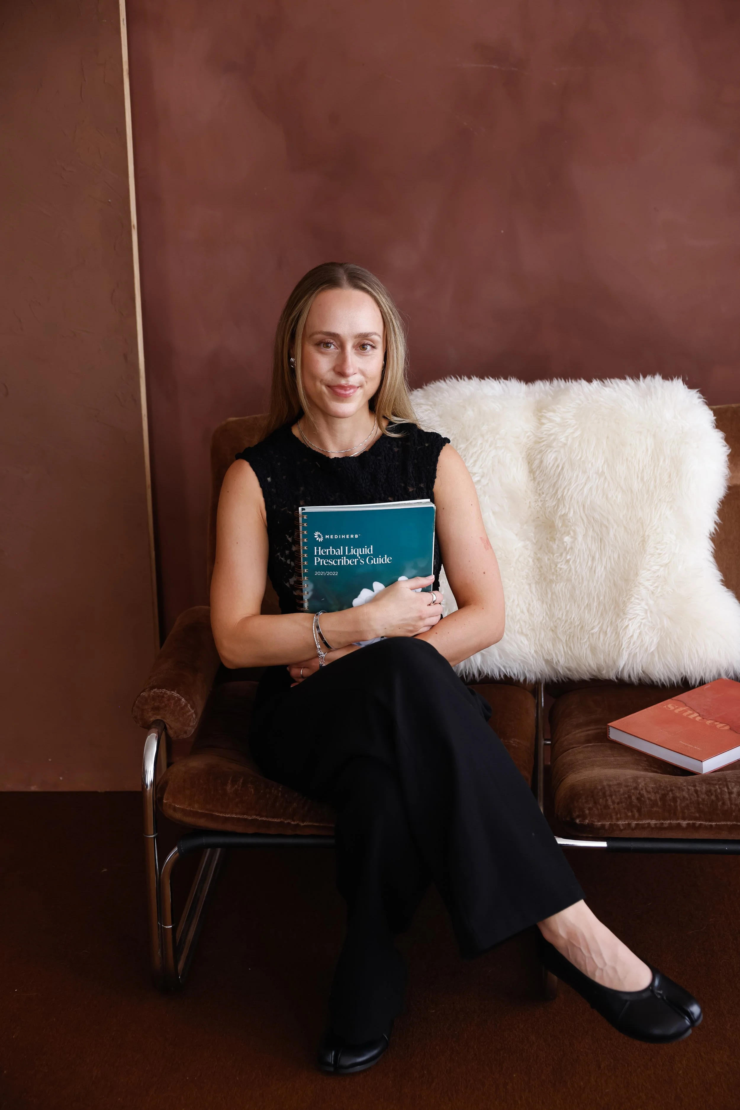 A young woman with long blonde hair sitting on a brown velvet chair, holding a blue pamphlet titled 'Herbal Liquid Prescriber's Guide'. She is wearing a black lace sleeveless top, black pants, and black shoes. There is a white furry pillow behind her on the chair and a closed orange book on the sofa to her right, all against a brown wall.