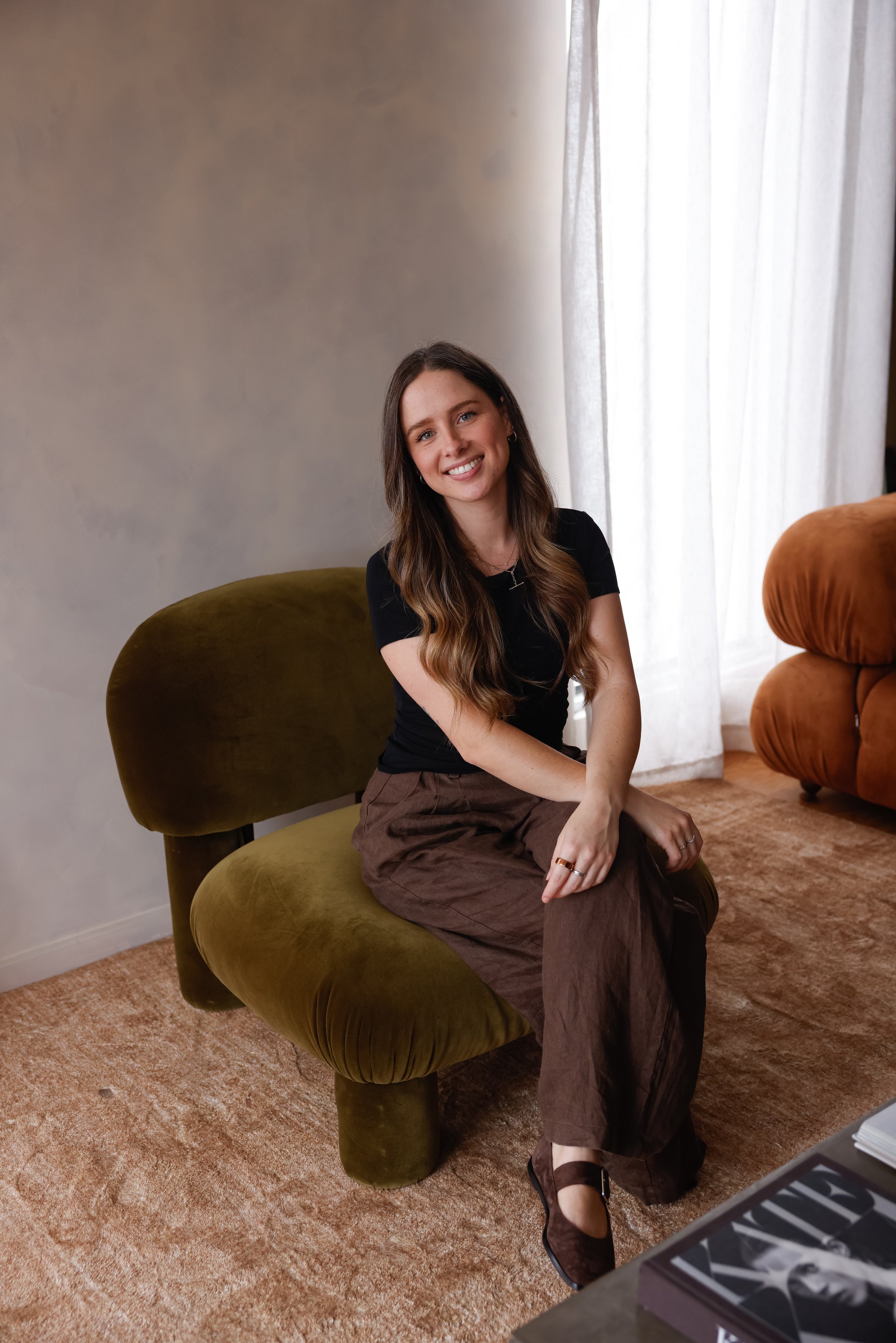A young woman with long brown hair and a black top sitting on an olive-green velvet chair in a room with beige carpet and white curtains, smiling at the camera.