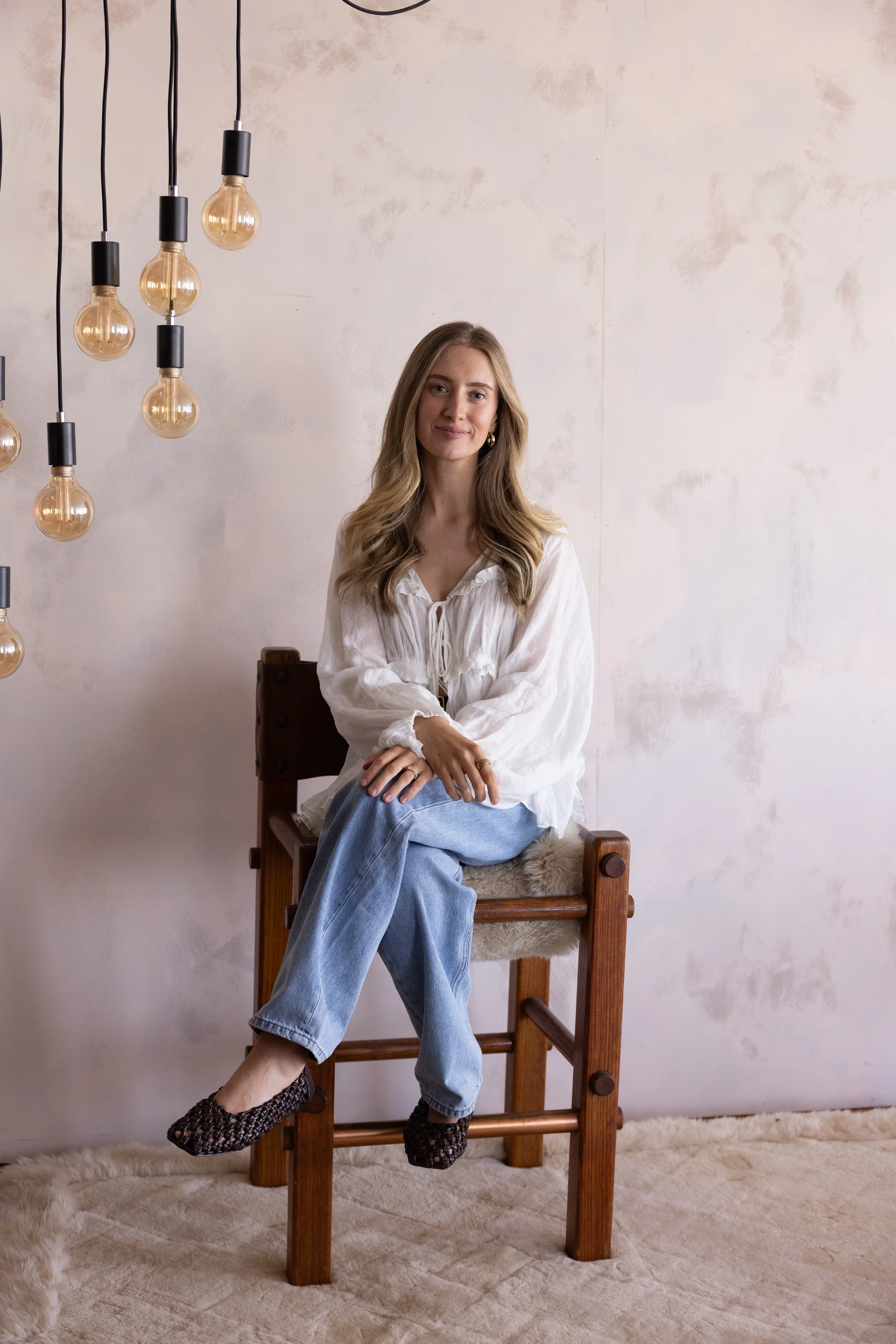 A young woman with long blond hair sitting on a wooden chair with a furry cushion, wearing a white blouse, jeans, and woven slip-on shoes, smiling slightly at the camera. There are hanging light bulbs on the left side and a plain wall in the background.