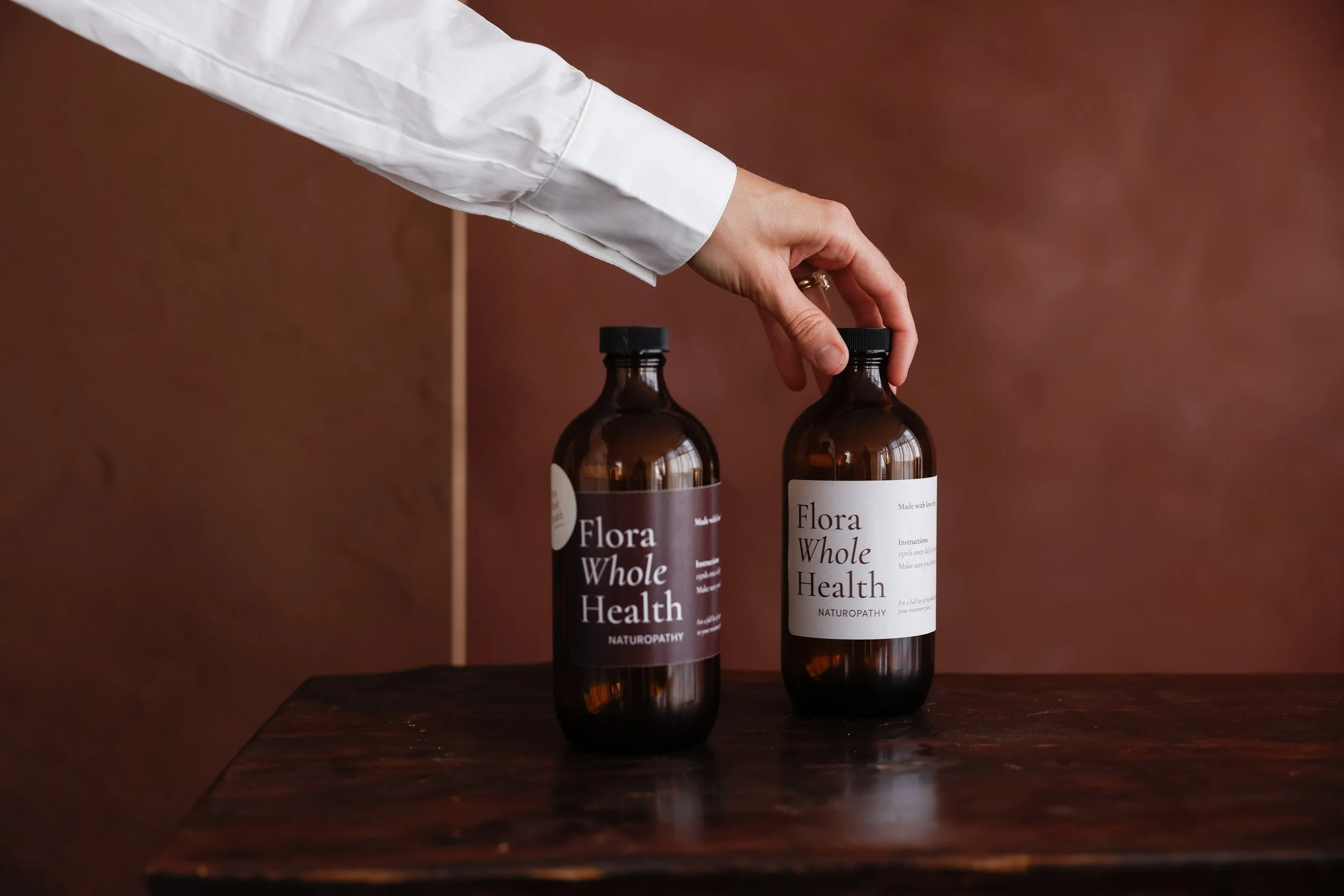 Two brown glass bottles with black caps placed on a dark wooden surface. The bottles have white labels with black text that reads "Flora Whole Health" and "Naturopathy." A person's hand, wearing a white long-sleeve shirt, is reaching towards the bottles. The background features a warm, reddish-brown wall.