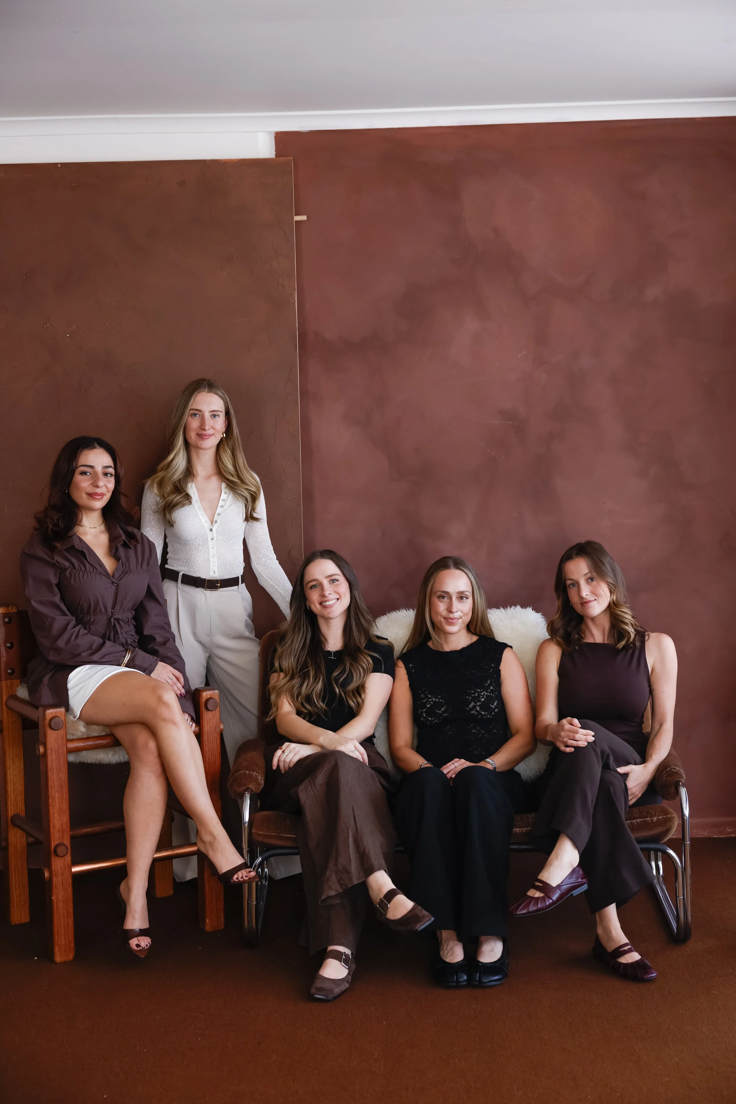 A group of six women with light to medium skin tones sitting on and standing around a vintage brown velvet sofa and chairs in a room with a brown textured wall. They are dressed in semi-formal attire and are smiling at the camera.