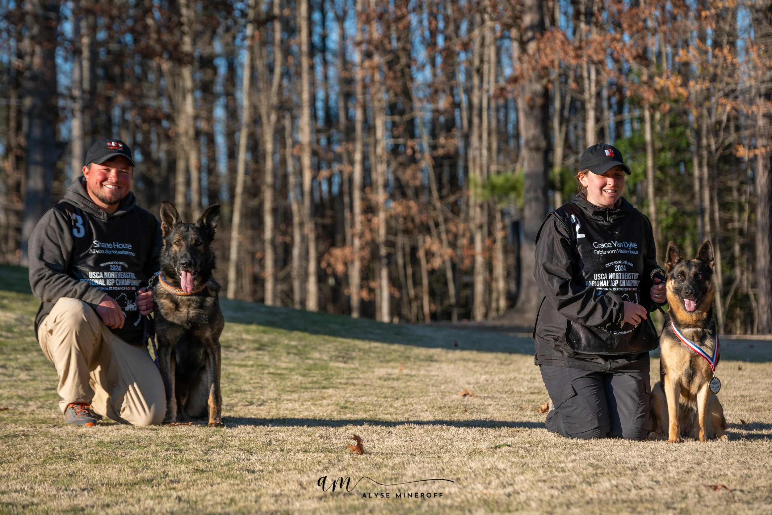 Two people kneeling on grass with their dogs, both dogs wearing medals, in a park with trees in the background.