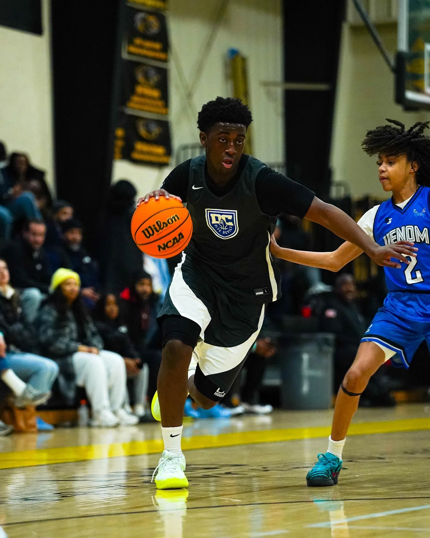 A young male basketball player in black and white uniform dribbling a basketball during a game, with an opponent in a blue jersey guarding him.