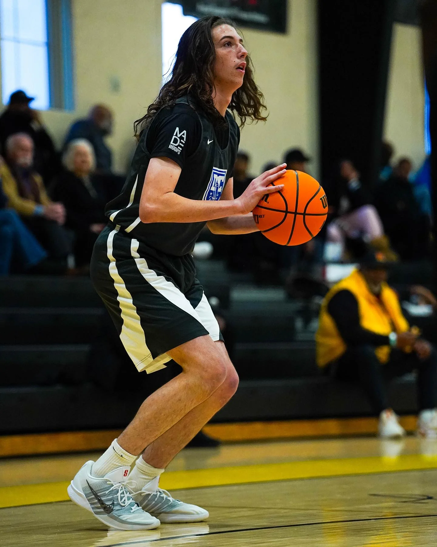 A basketball player holding a basketball on a court, preparing to shoot, with spectators seated in the background.