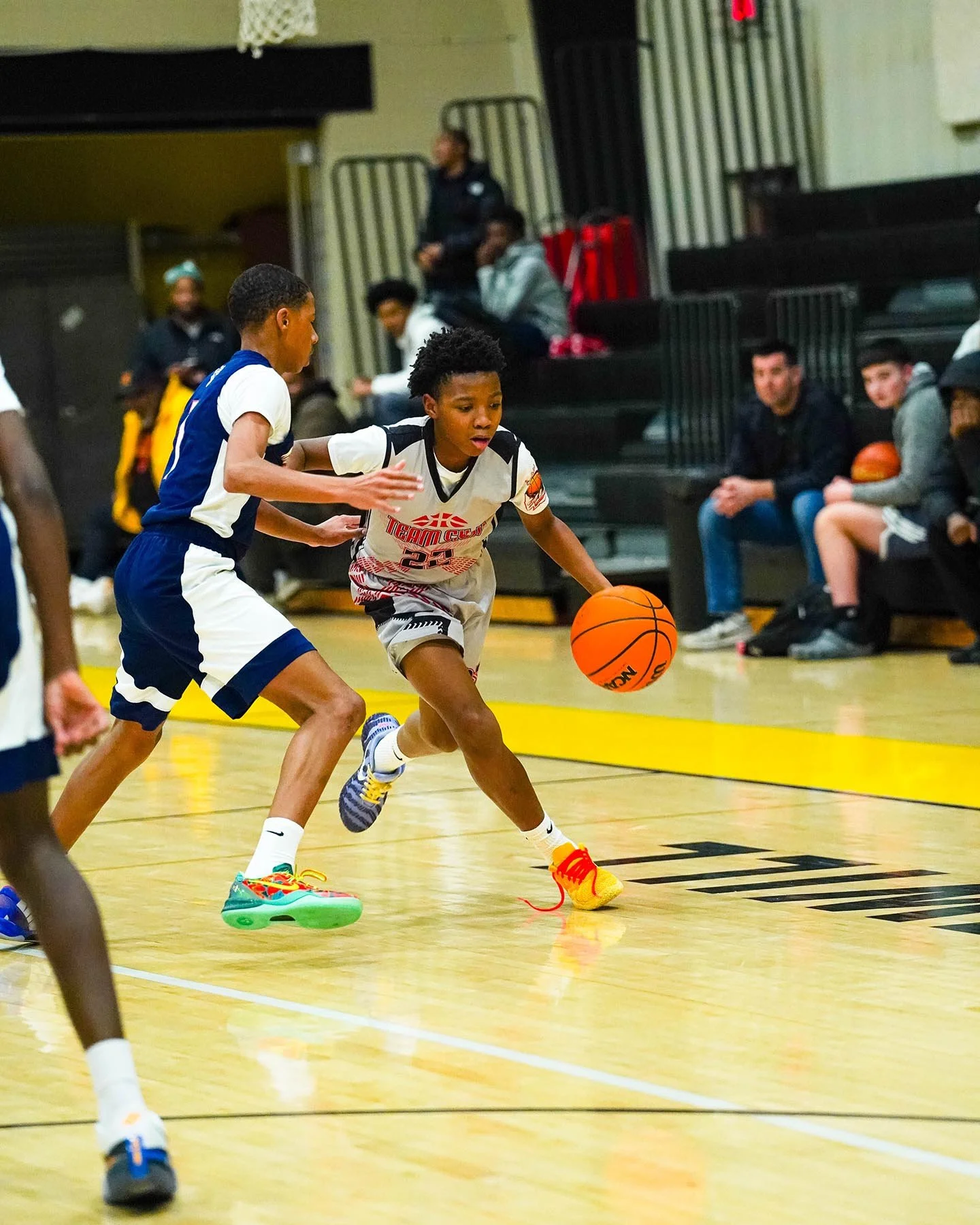 Two young boys playing basketball in an indoor gym, one in a white jersey dribbling the ball while the other in a blue and white jersey defends.