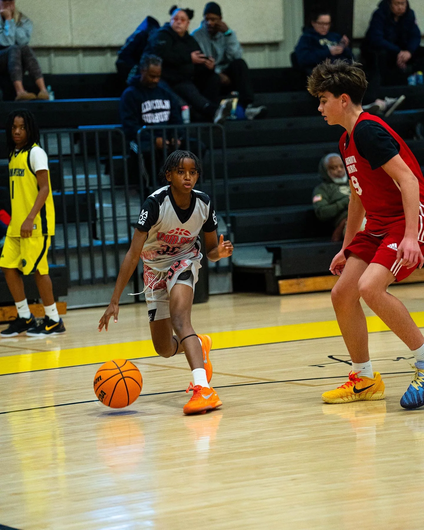 Young basketball player dribbling on indoor court during a game, with other players and spectators in the background.