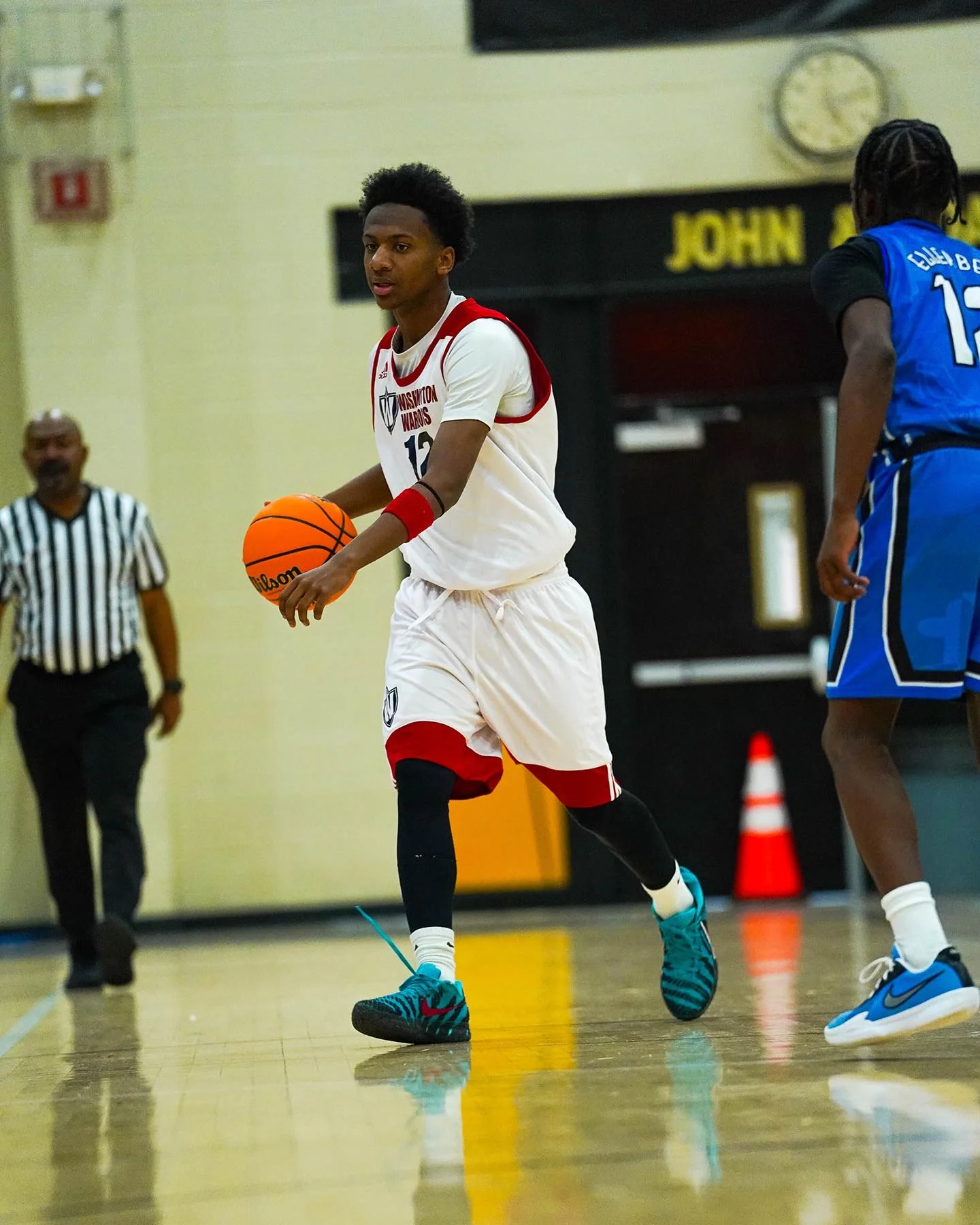 A young male basketball player wearing a white and red Washington Warriors uniform dribbling a basketball on an indoor court. Another player in a blue uniform is partially visible to his right, and a referee is in the background.