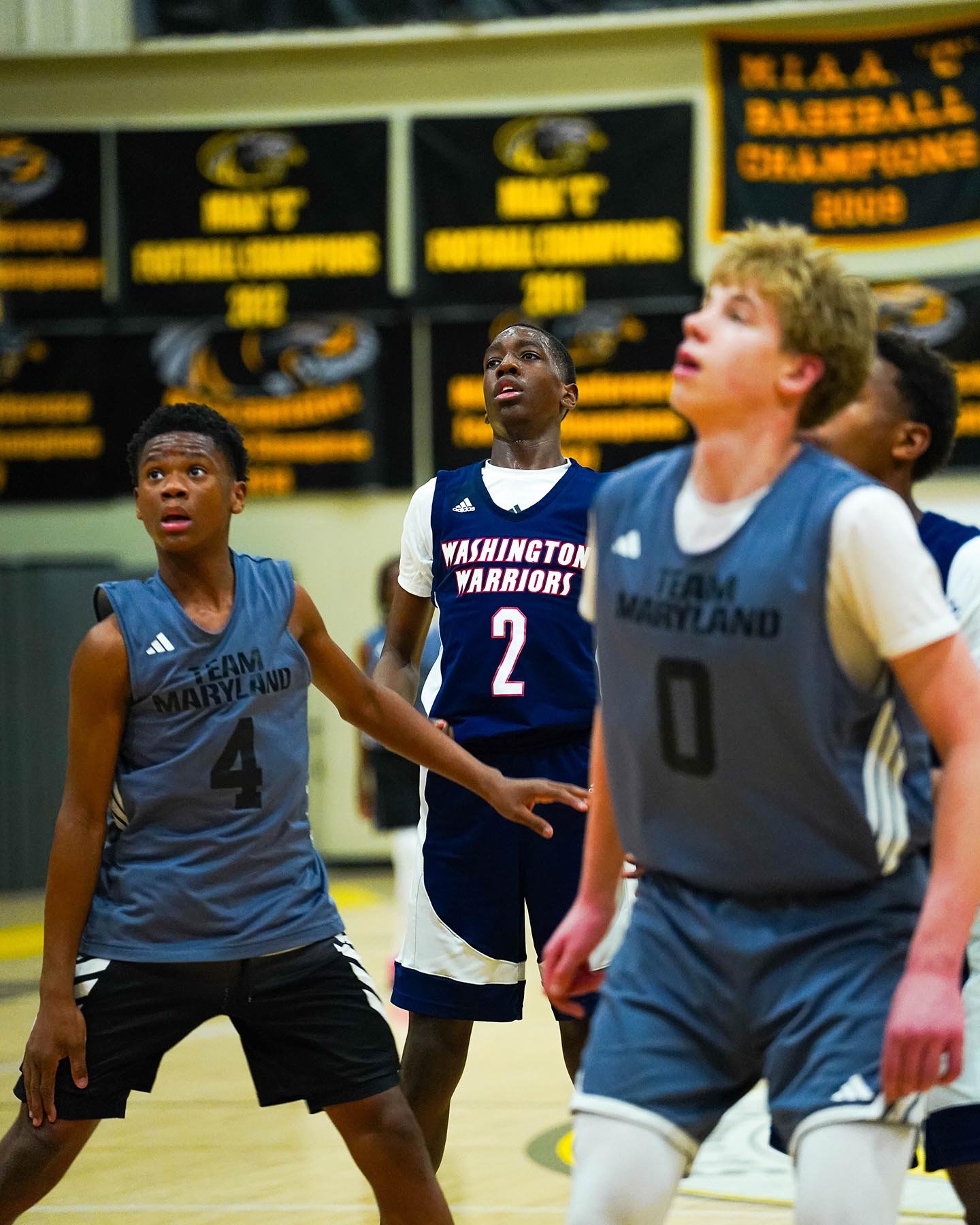 Young boys playing basketball, wearing team jerseys for Team Maryland and Washington Warriors, in a gymnasium with banners in the background.
