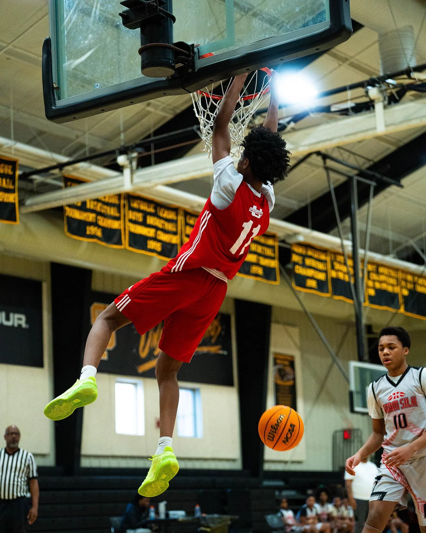 A young basketball player in a red and white uniform is hanging from the basketball hoop, about to score. His game is being played in an indoor gymnasium with banners hanging from the ceiling and some spectators and officials in the background.