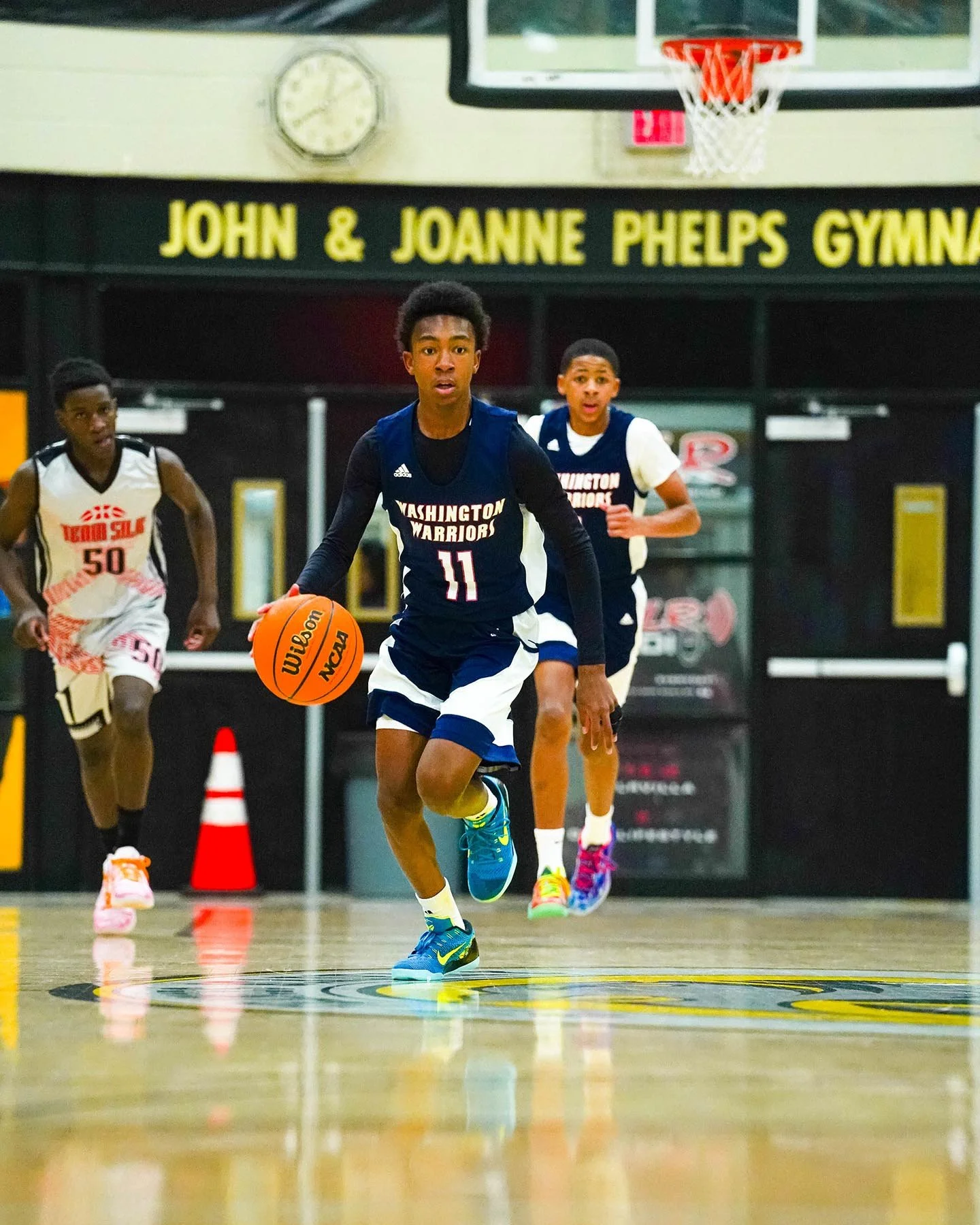 Young basketball player in Washington Warriors uniform dribbling a basketball on court, with two other players in background, inside John & Joanne Phelps Gymnasium.