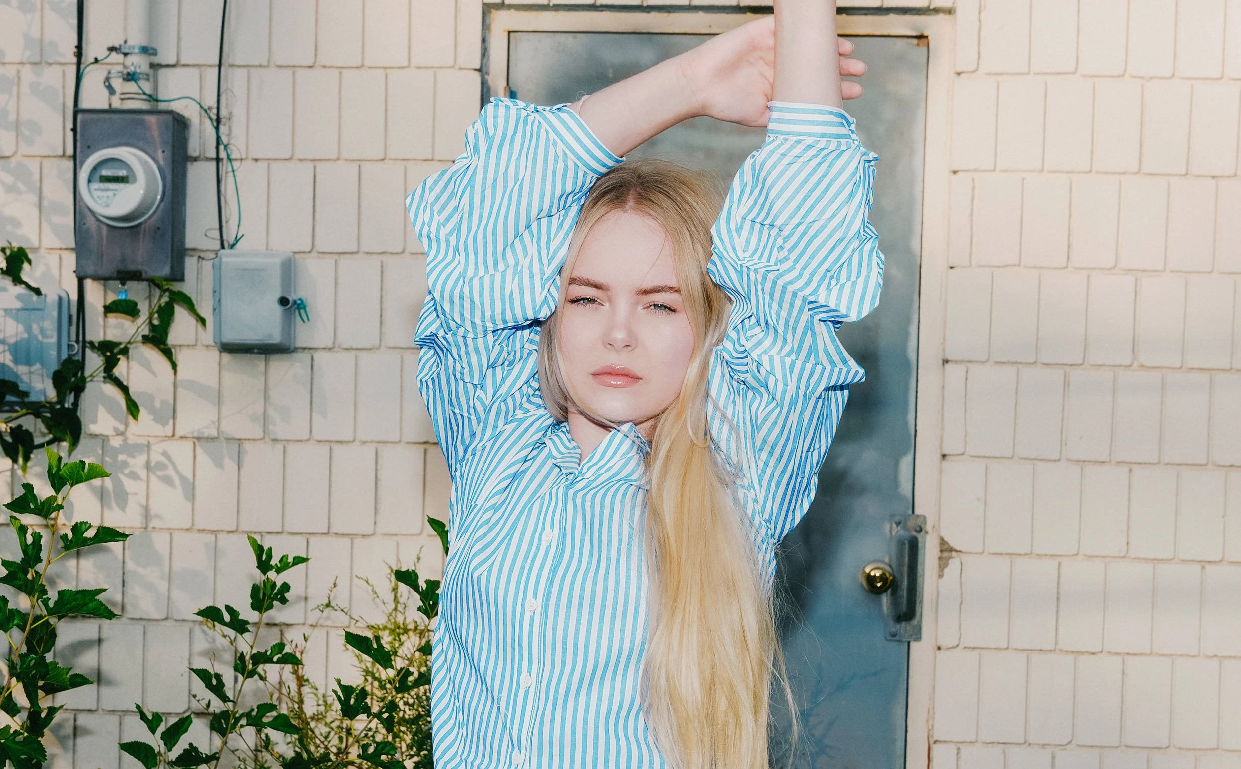Blonde woman in a blue-and-white striped shirt posing with her arms raised in front of a tiled wall and blue door
