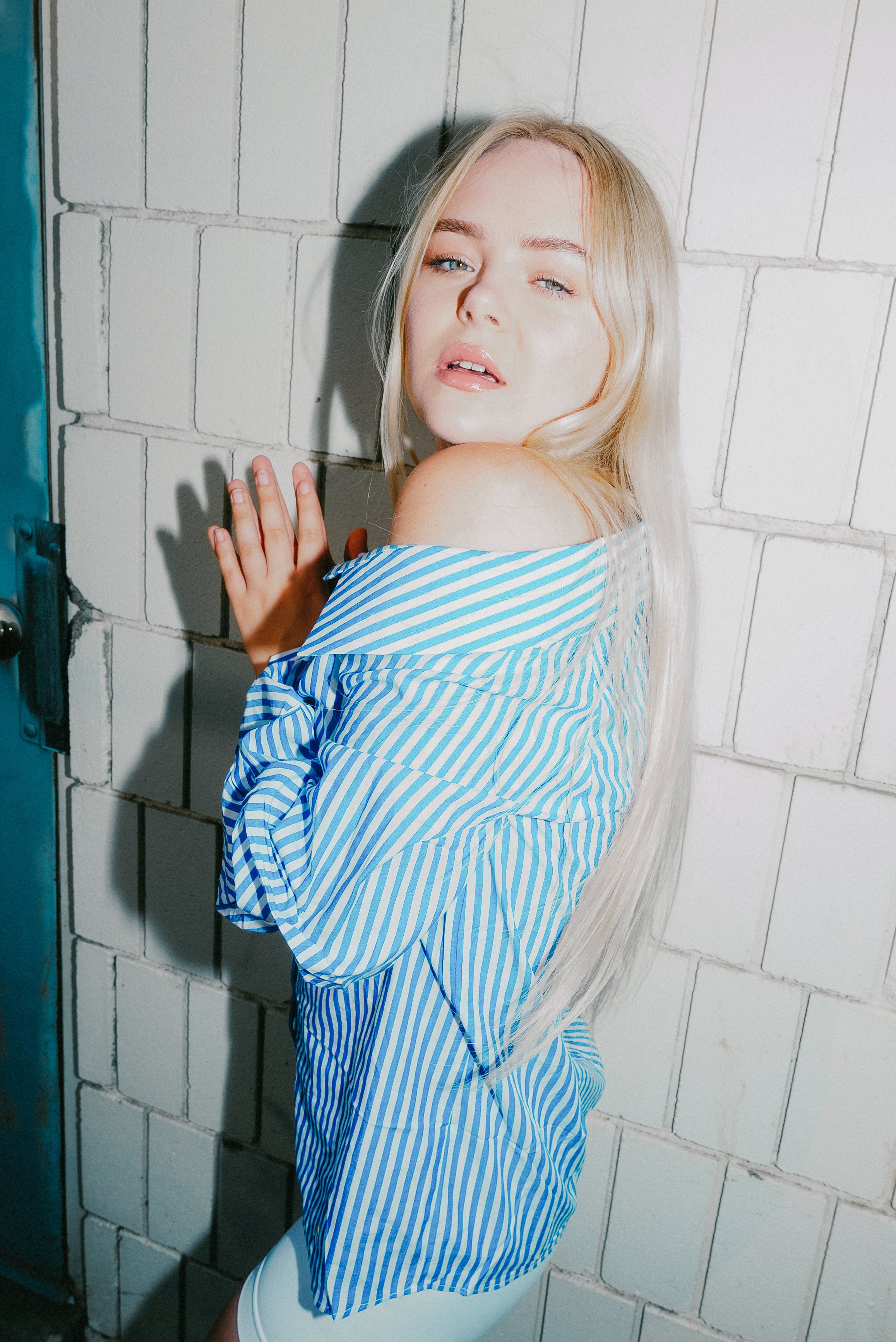 Blonde woman in a blue-and-white striped top posing against a white brick wall