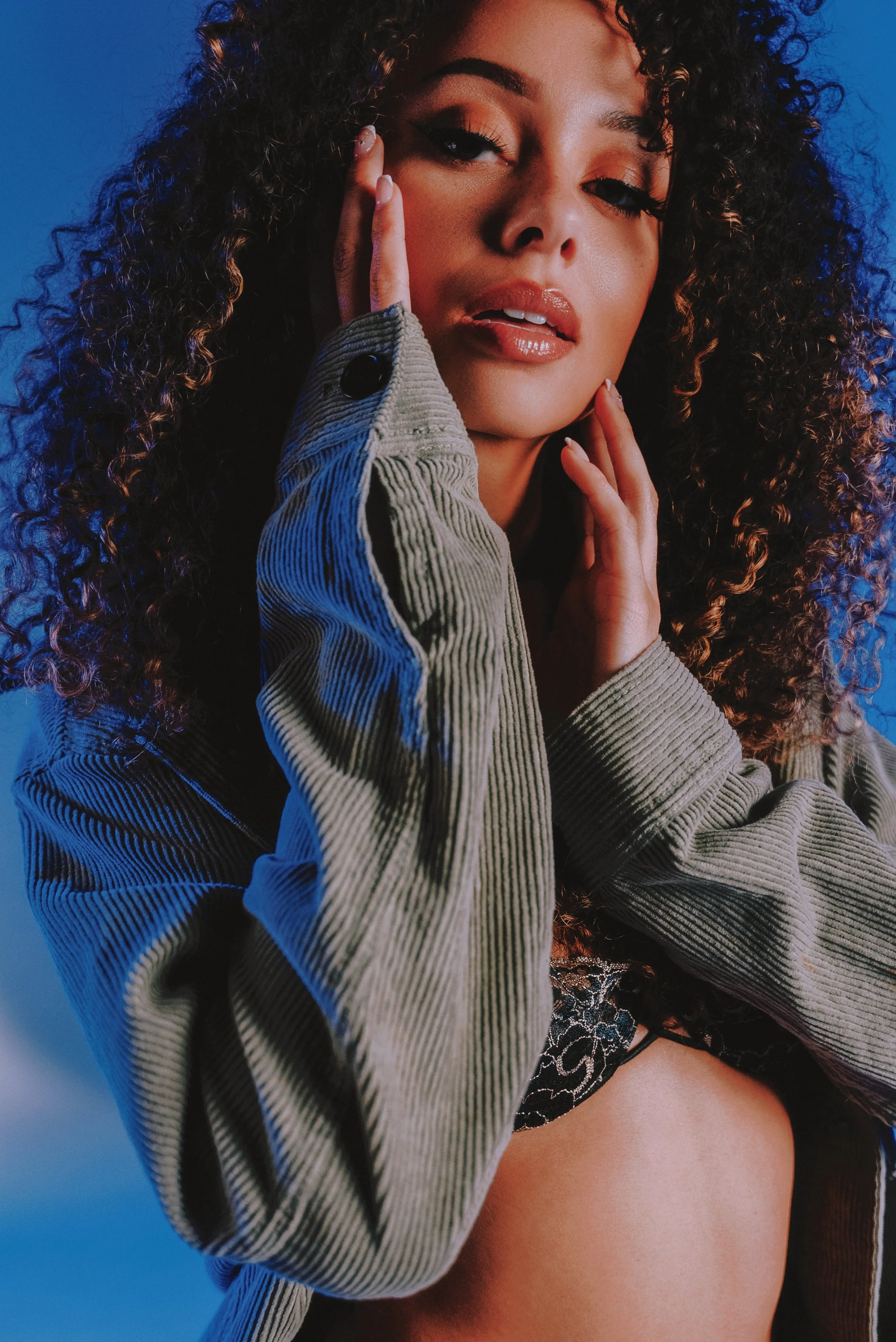 Close-up portrait of a curly-haired woman with dramatic makeup and manicured fingers against a vivid blue background.