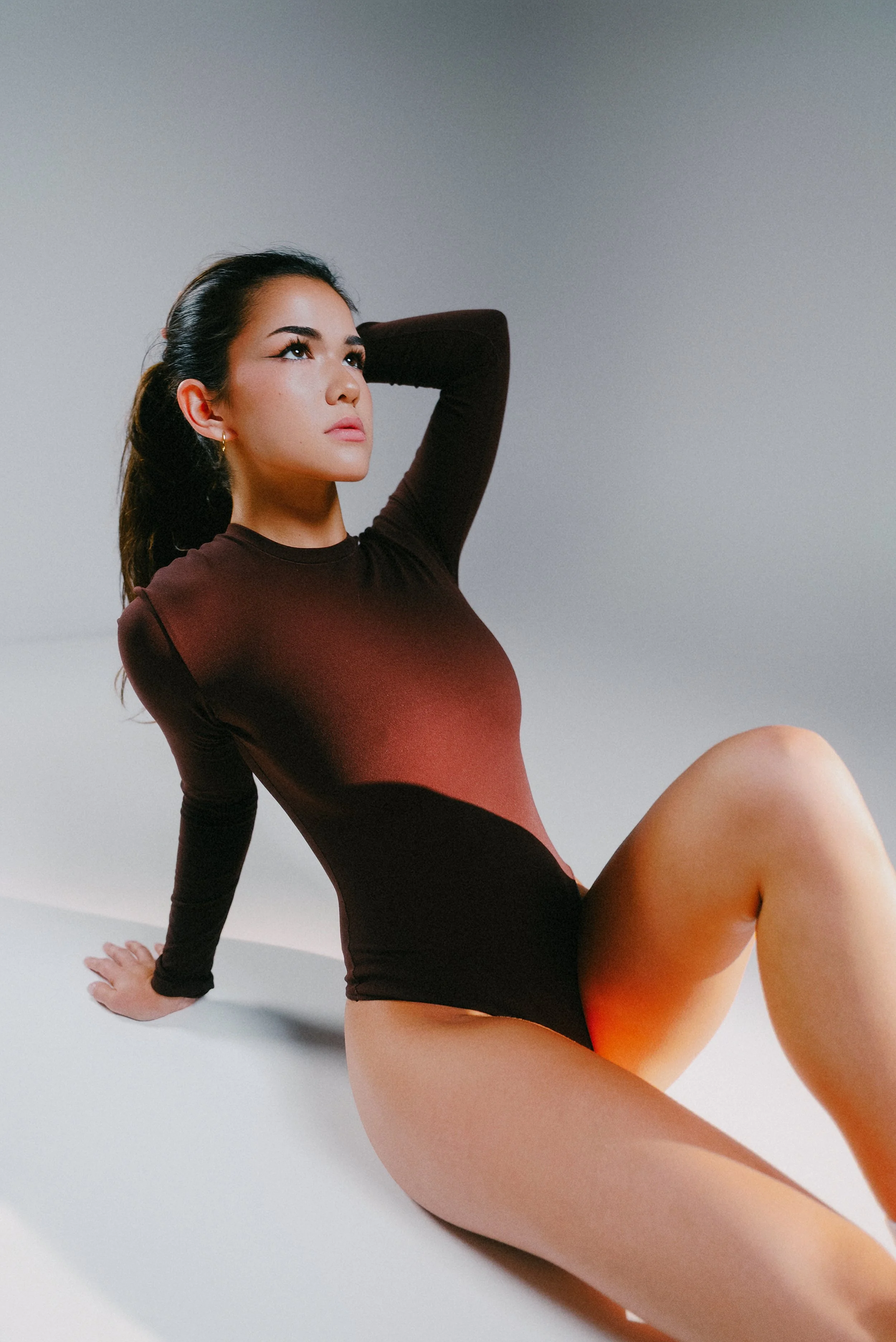 Woman in a dark long-sleeve bodysuit reclining on a white floor with one arm behind her head