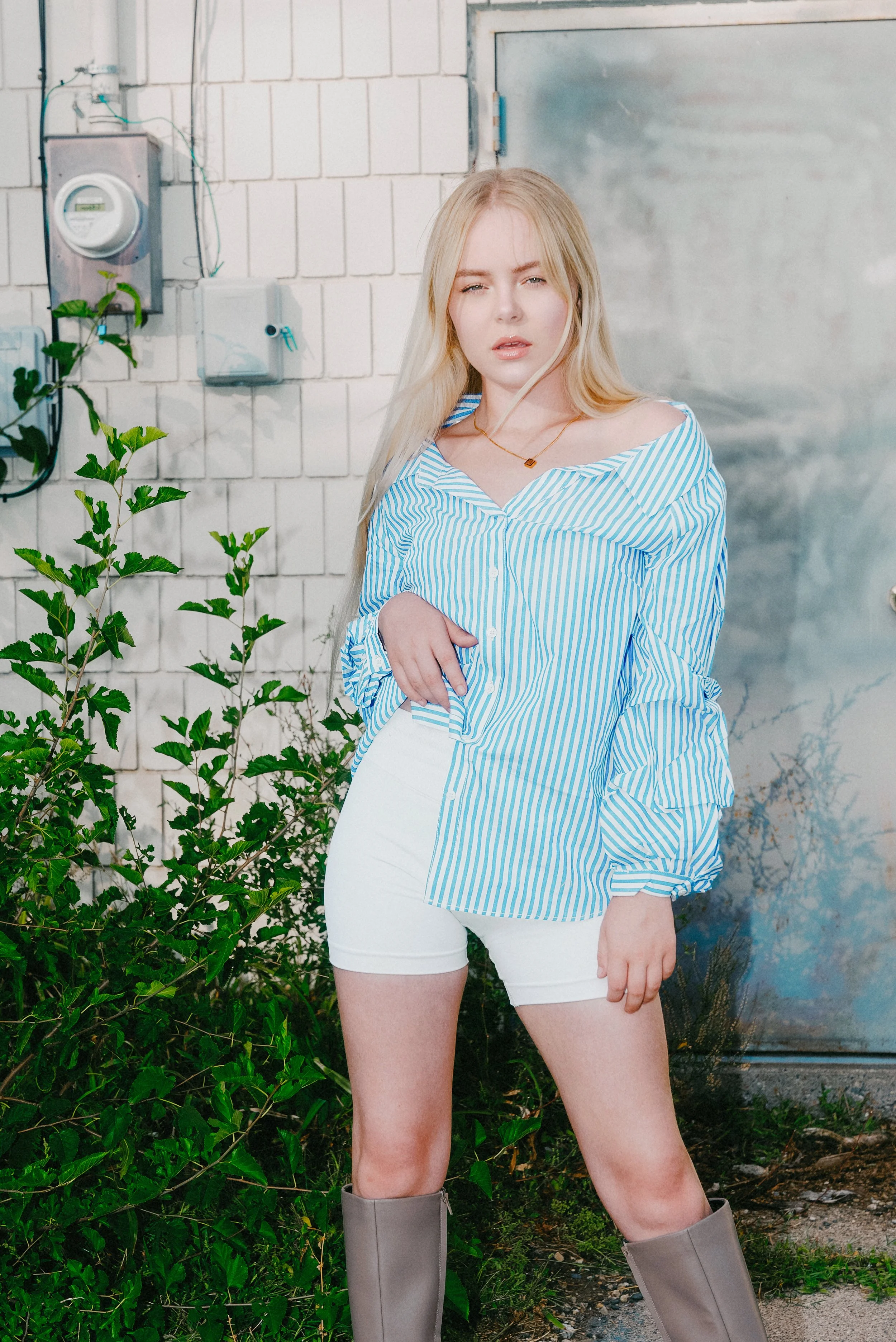 Blonde woman in a blue-and-white striped shirt, white shorts, and knee-high boots standing beside greenery and a white brick wall