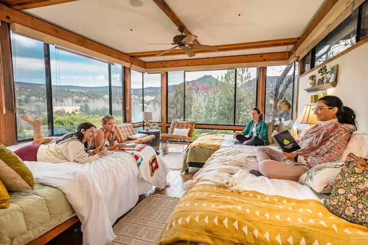 A spacious sunroom with large glass windows showcasing a scenic view of trees and mountains. Inside, four women are relaxing on beds and lounge chairs, some reading or using laptops, with natural light filling the space.