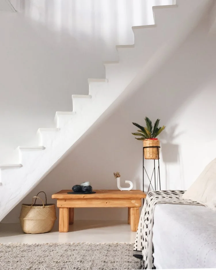 Minimalist living room with a white wall, staircase, wooden coffee table, woven basket, potted plant on a stand, and a sofa with a blanket.