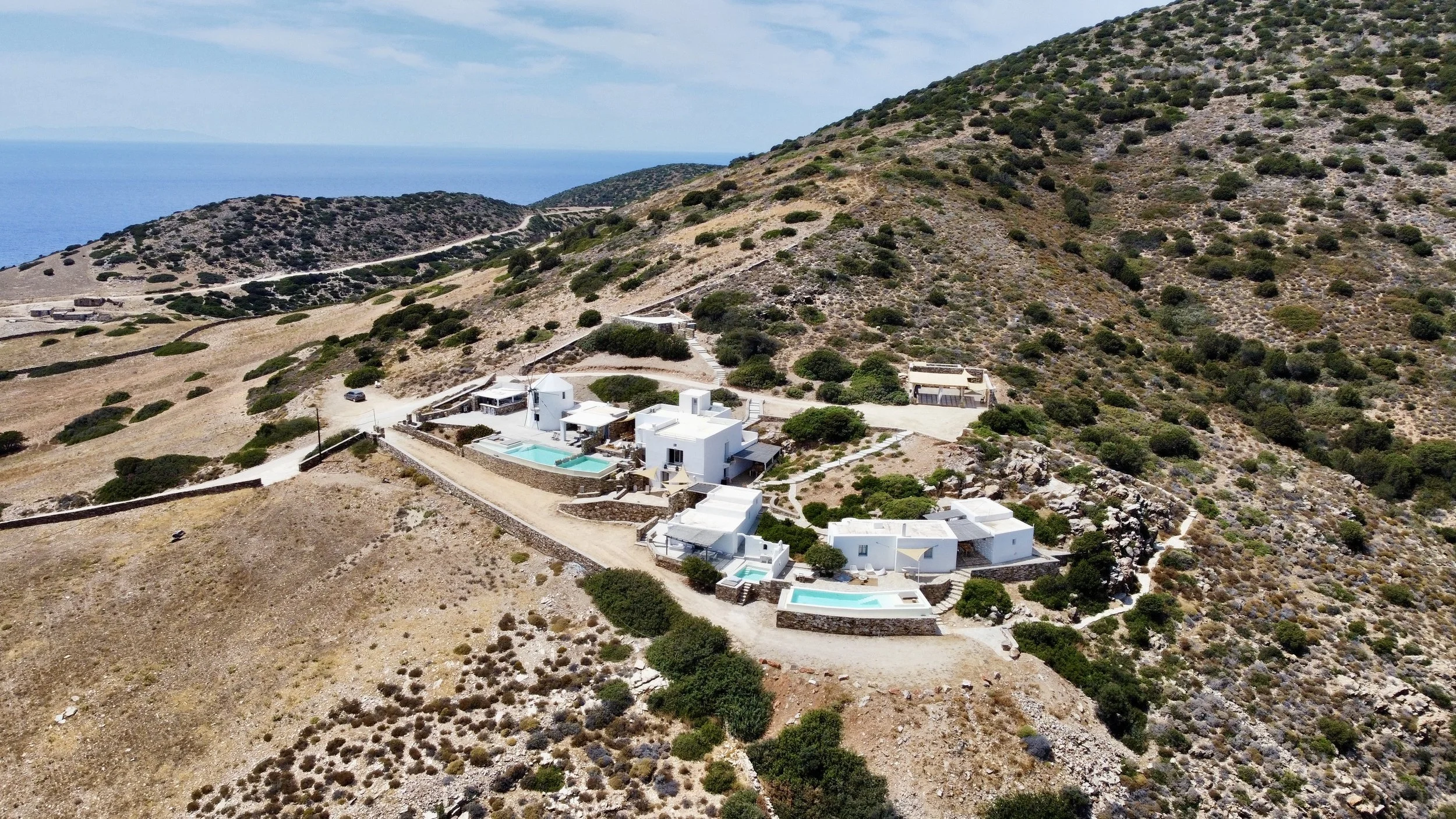 Aerial view of a white hillside villa complex with multiple pools, situated on a dry, rocky hillside with sparse greenery, overlooking the sea in the background.