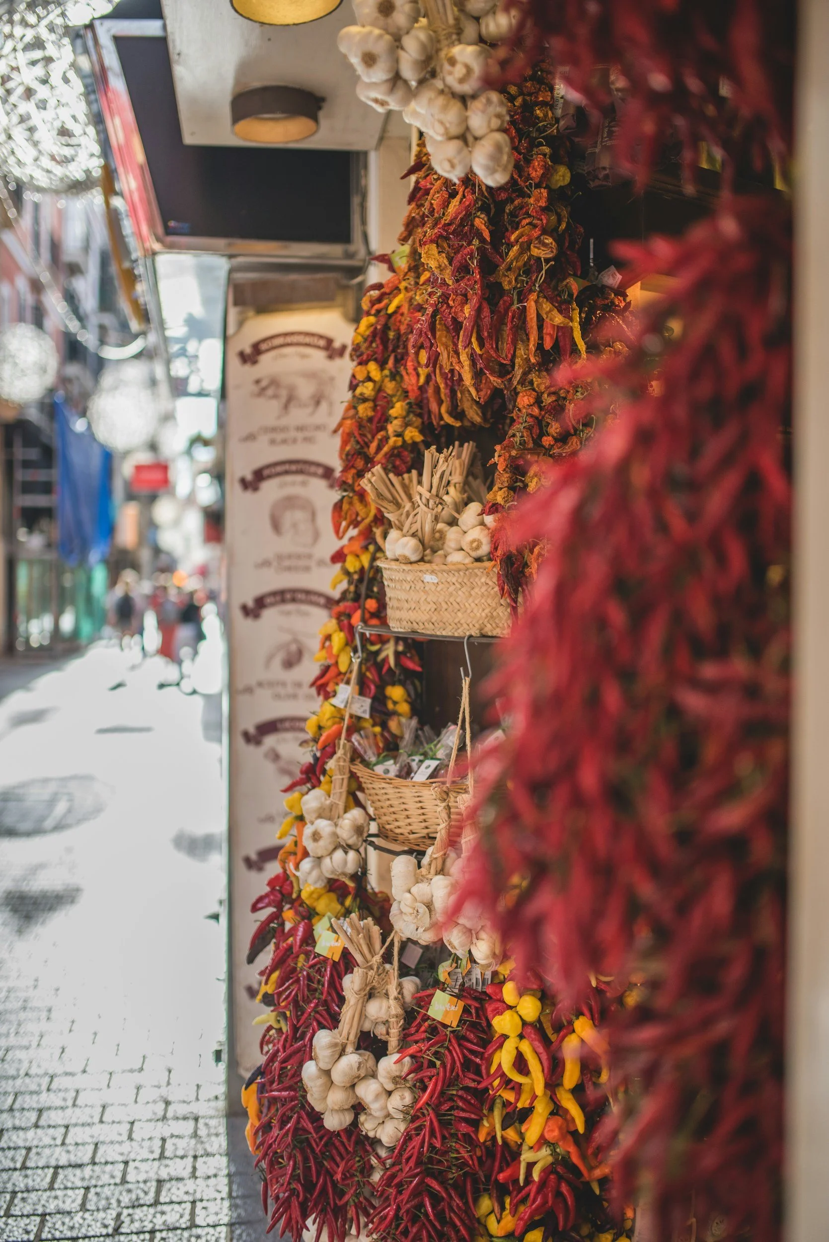 Display of garlic braids and colorful chili peppers at a street market stall, with a blurred pedestrian and urban street scene in the background.