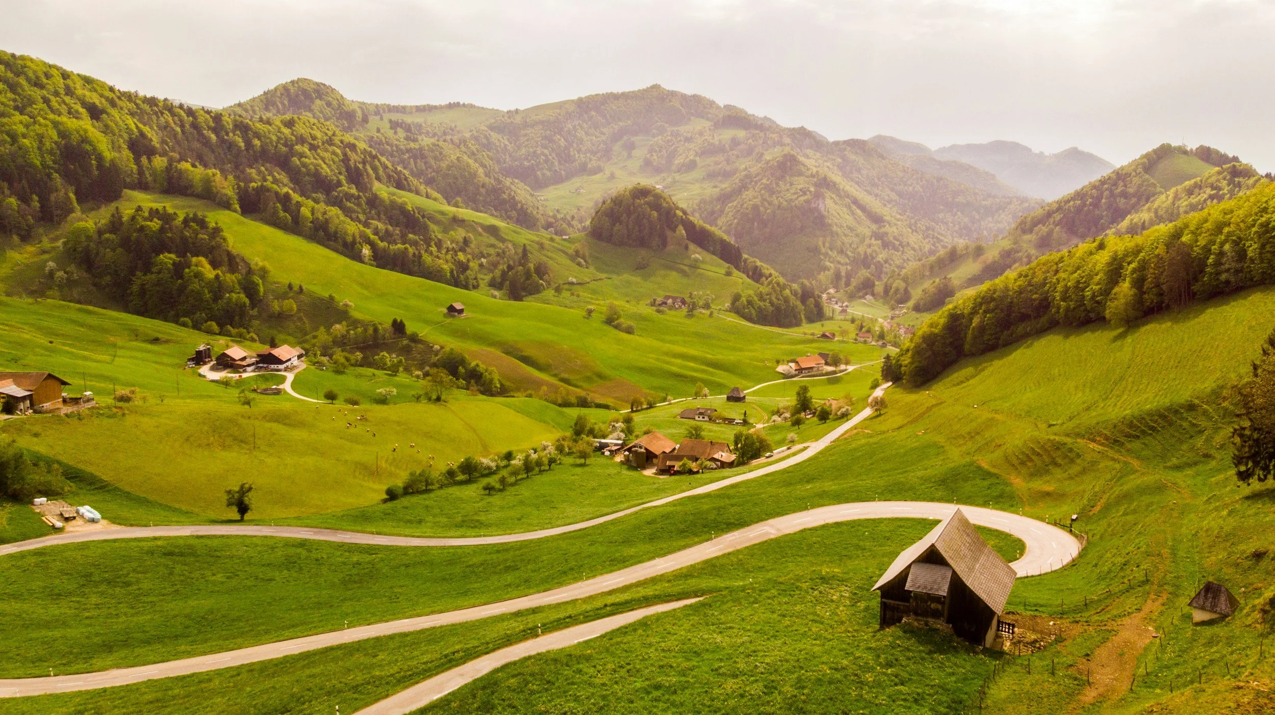 Scenic view of green rolling hills with small houses and winding roads, surrounded by forested mountains.