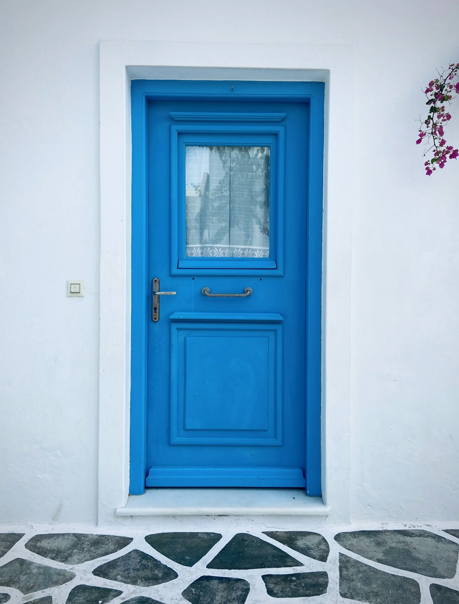 Blue door with a window, set in a white wall, with a small step at the bottom and a patterned stone pathway in front.