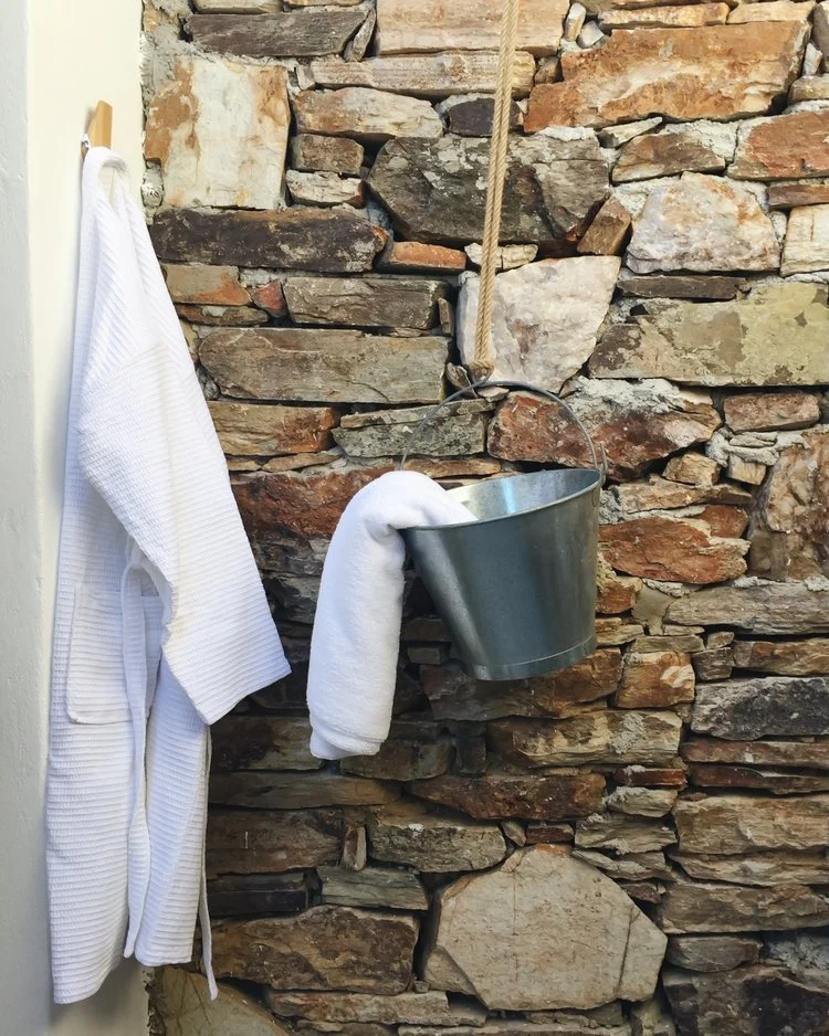 A white bathrobe hanging on a hook, a white towel hanging over the side of a metal bucket, which is suspended on a wall made of irregularly shaped stones by a rope.