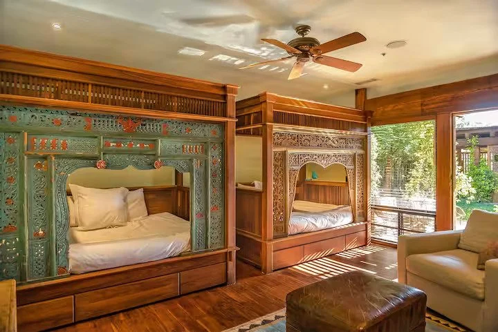 A cozy bedroom with two wooden canopy beds featuring intricate carvings, a ceiling fan, a beige armchair, a brown ottoman, and large windows with greenery outside.