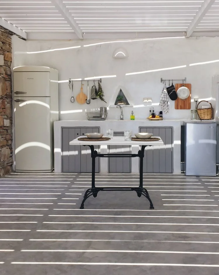 Sunlight filtering through a slatted roof creates shadow lines across an outdoor kitchen with a white and gray color scheme, featuring a vintage refrigerator, a sink with hanging utensils, small countertop appliances, and a table set for two.