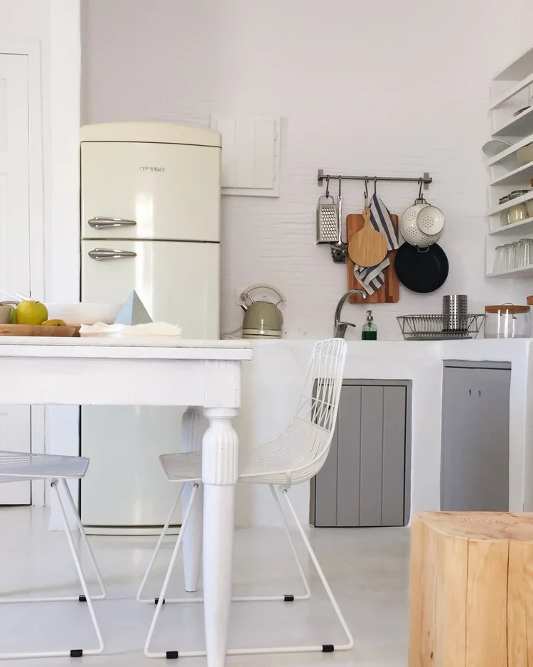 A minimalist, white kitchen with a vintage cream-colored refrigerator, white table, and wire chairs. The kitchen features hanging pots, pans, and kitchen tools on a rail, with open shelving holding dishes and glassware.