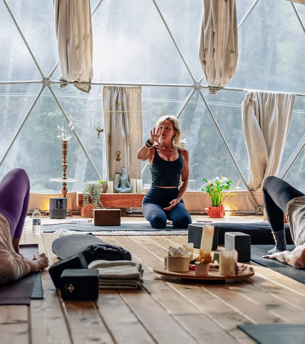 Woman leading a yoga class in a dome-shaped room with natural light, surrounded by yoga mats, candles, a plant, and decorative items.