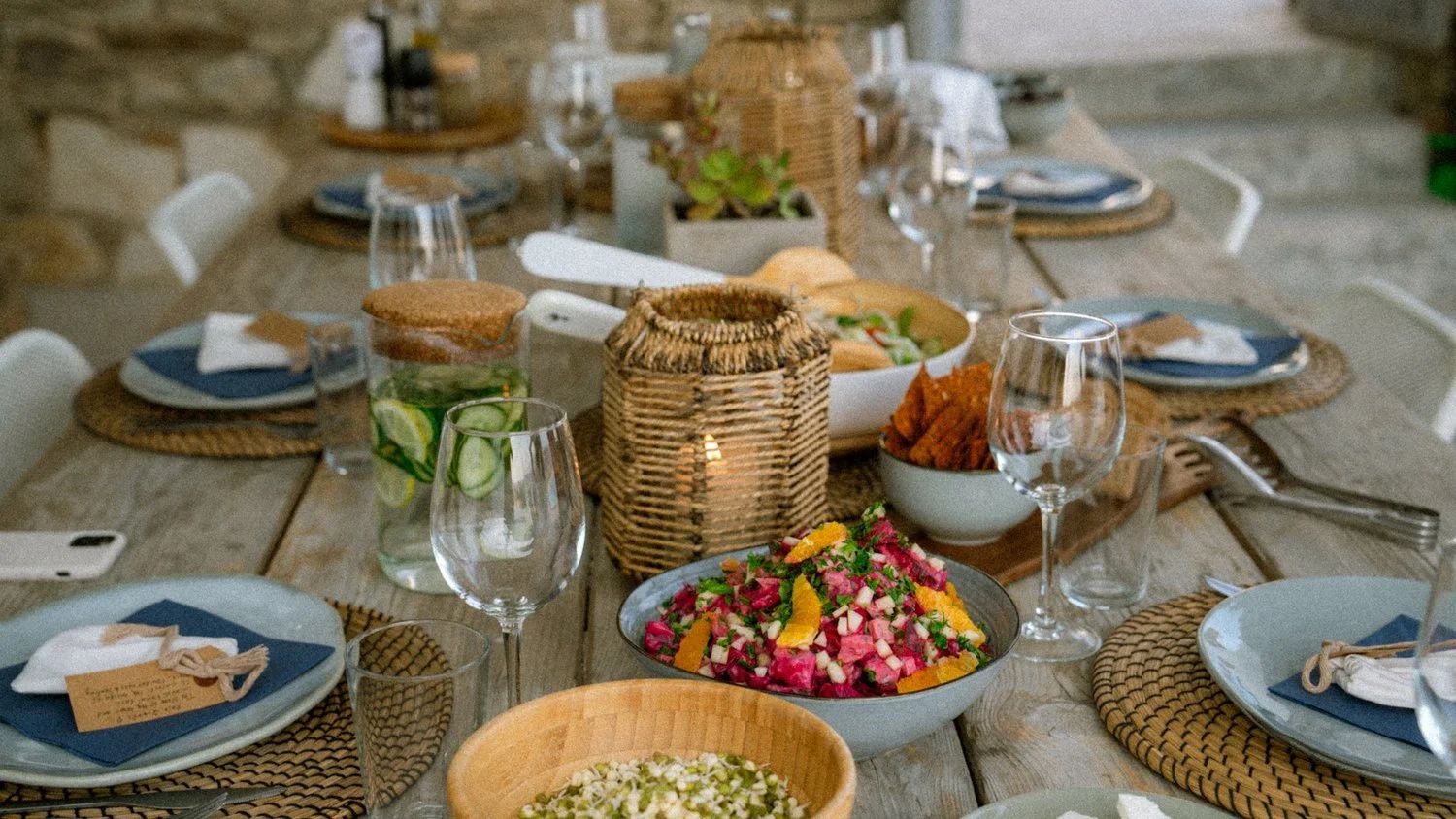 A rustic dining table set with plates, wine glasses, and a variety of colorful salads and appetizers, with a candle in a woven basket at the center.