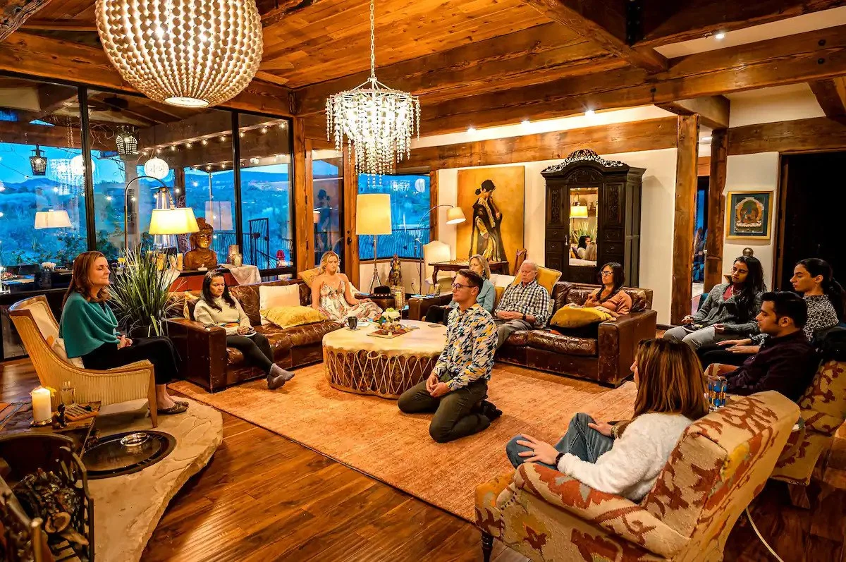 A group of people gathered in a cozy, warmly lit living room with wooden beams and large windows. They are seated on various chairs and sofas, listening attentively to a woman speaking. The room features elegant lighting fixtures, artwork on the walls, and a mixture of vintage and modern decor.