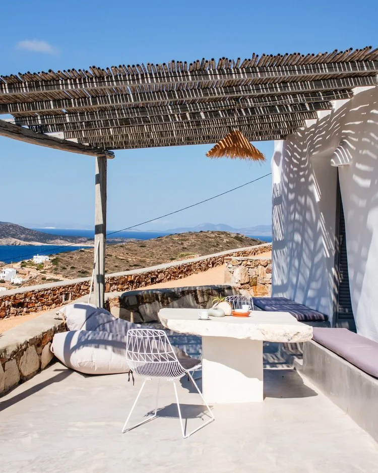 Outdoor seating area with a white marble table, wire chair, and cushioned bench under a wooden pergola. Coastal landscape with blue sky, sea, and hills in the background.