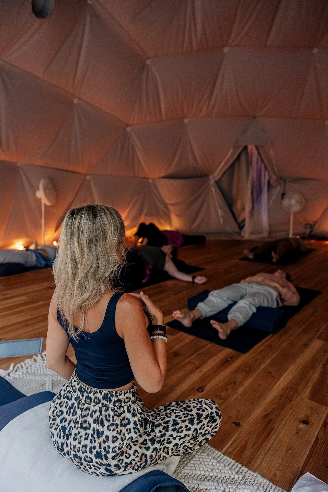A woman practicing yoga or meditation in a dimly lit, cozy indoor space with a wooden floor and fabric-covered walls, while others lie on mats in the background.