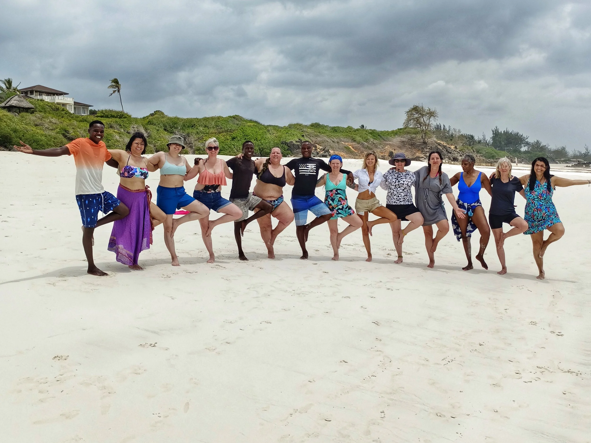 Group of fifteen people standing on a sandy beach, holding hands and raising their legs, with green hills, houses, and cloudy sky in the background.