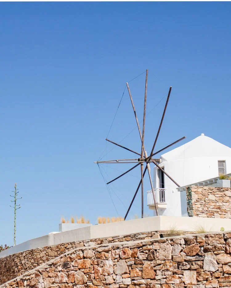 A white house with a stone wall and a large wooden windmill on the roof, set against a clear blue sky.