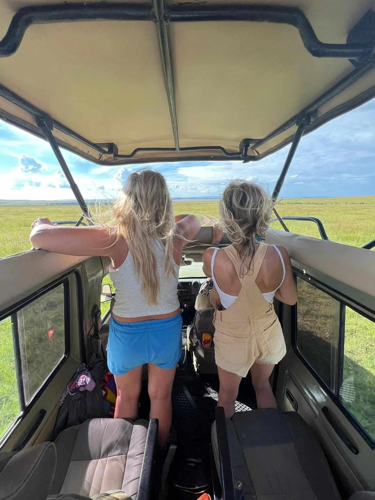 Two women with blonde hair inside a safari vehicle, looking out over a grassland landscape under a blue sky with clouds.