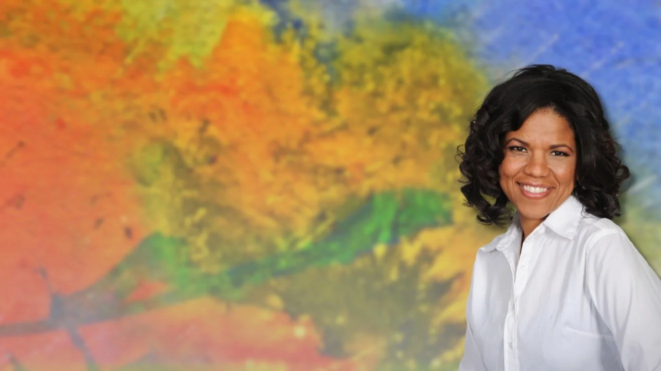 Smiling woman with dark curly hair wearing a white shirt, standing in front of a colorful, abstract background of orange, yellow, green, and blue hues.