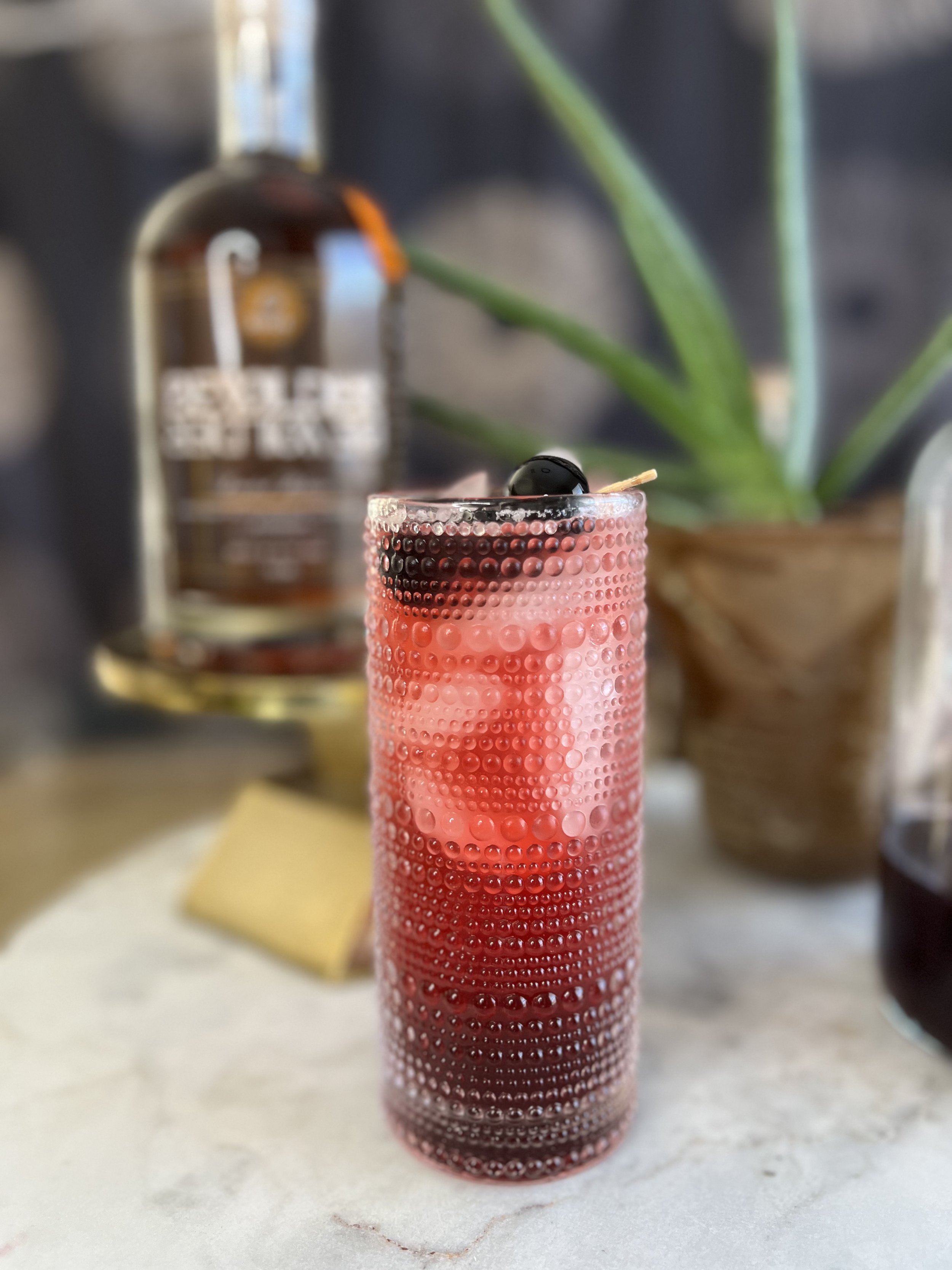 A tall pink cocktail glass with a textured dotted pattern, filled with ice and a dark cherry on top, placed on a light-colored surface.