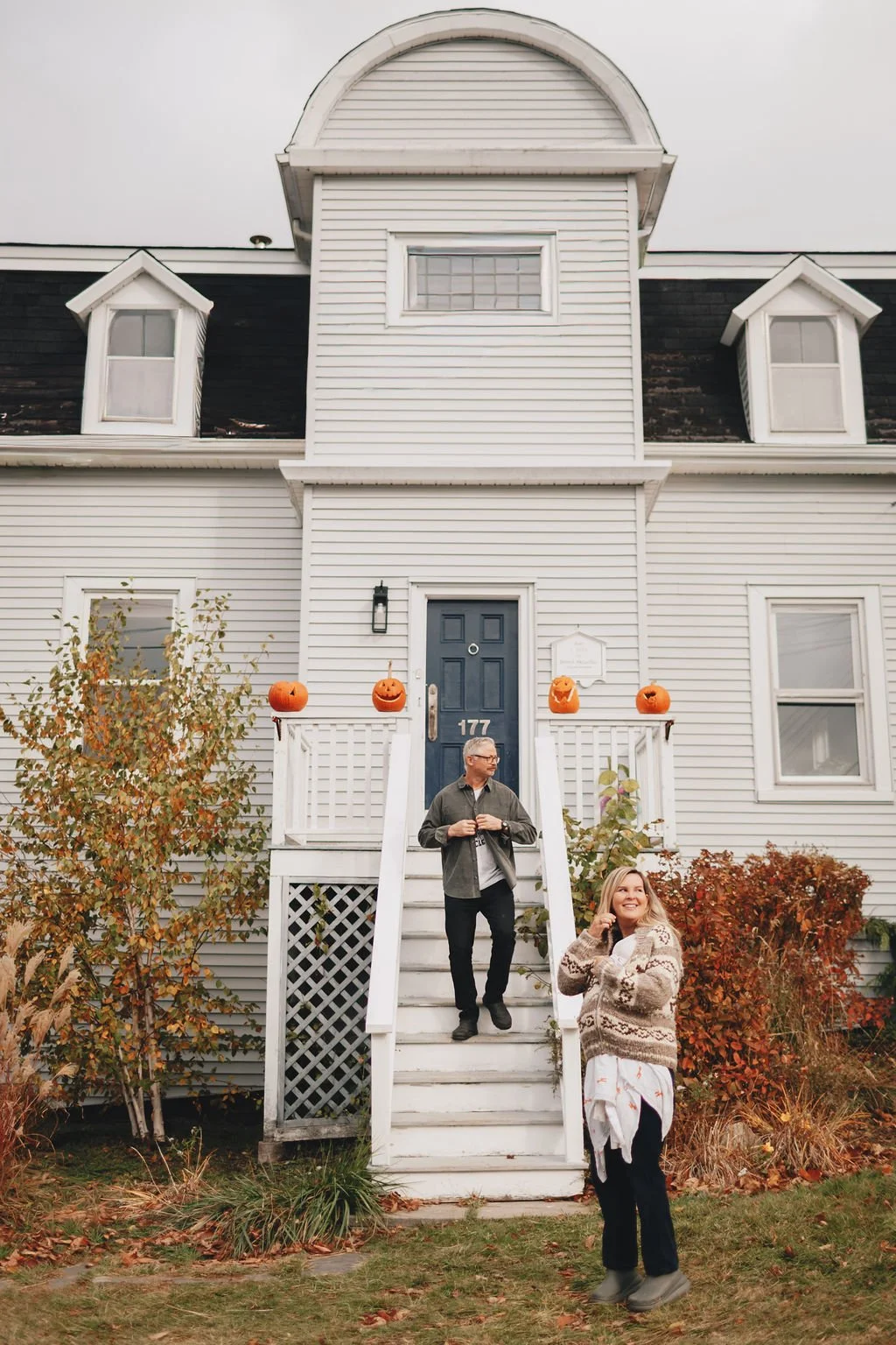 Two people standing on the front steps of a white house decorated with Halloween pumpkins. The house has a blue door, white siding, and dormer windows with black roofs. The man is on the stairs, looking to the side, while the woman is smiling, talking on a cell phone, and standing on the grass. Autumn foliage surrounds the scene.