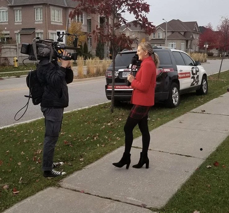 A woman in a red coat and black boots being interviewed on the sidewalk by a camera operator. A police vehicle with 'Breaking News' signage is parked behind her. Residential houses are visible in the background during fall.