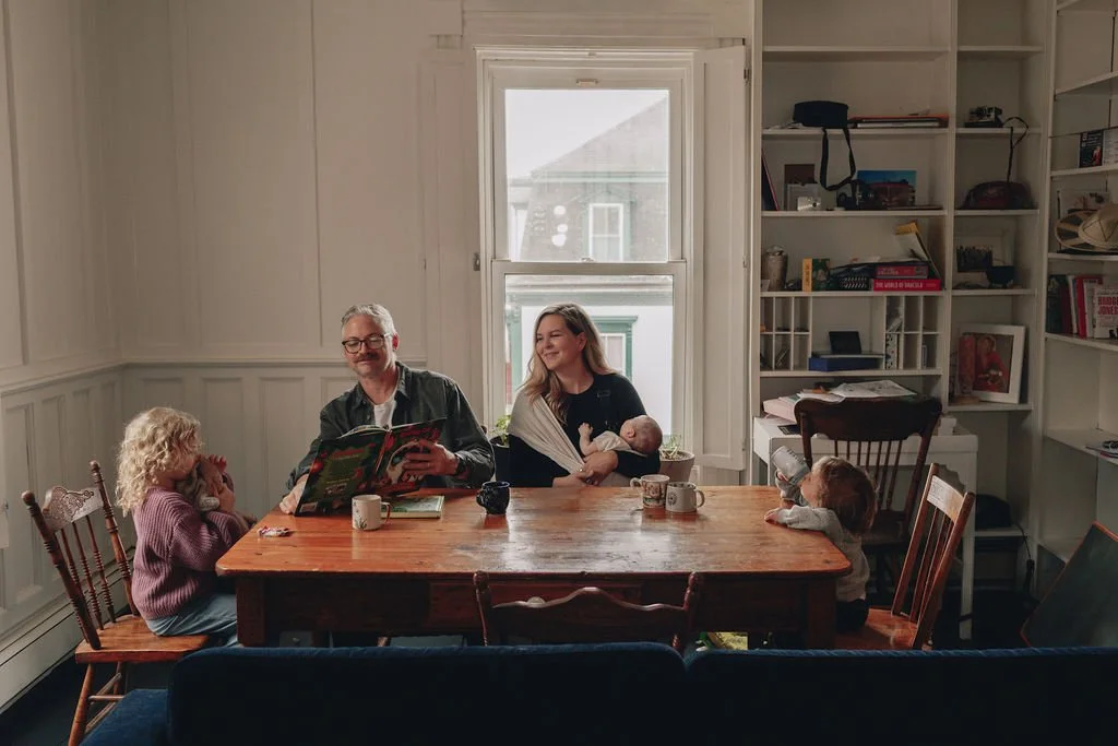 Family sitting at a wooden dining table in a room with white paneled walls and bookshelves. A woman holds a baby, a man reads a book, and two young girls sit at the table with mugs.