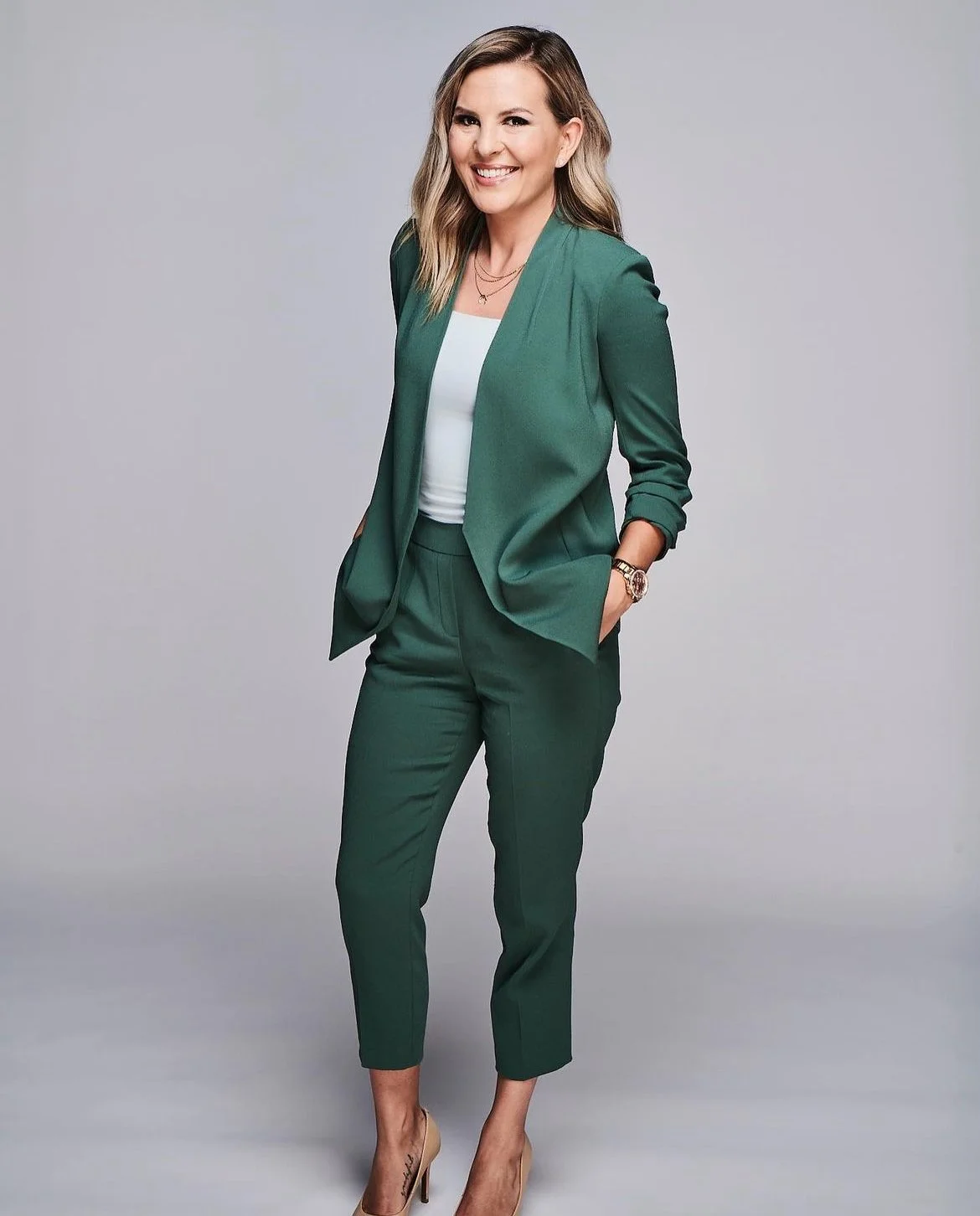 A woman in a green pantsuit with a white top, posing with one hand in her pocket, smiling in front of a neutral gray background.
