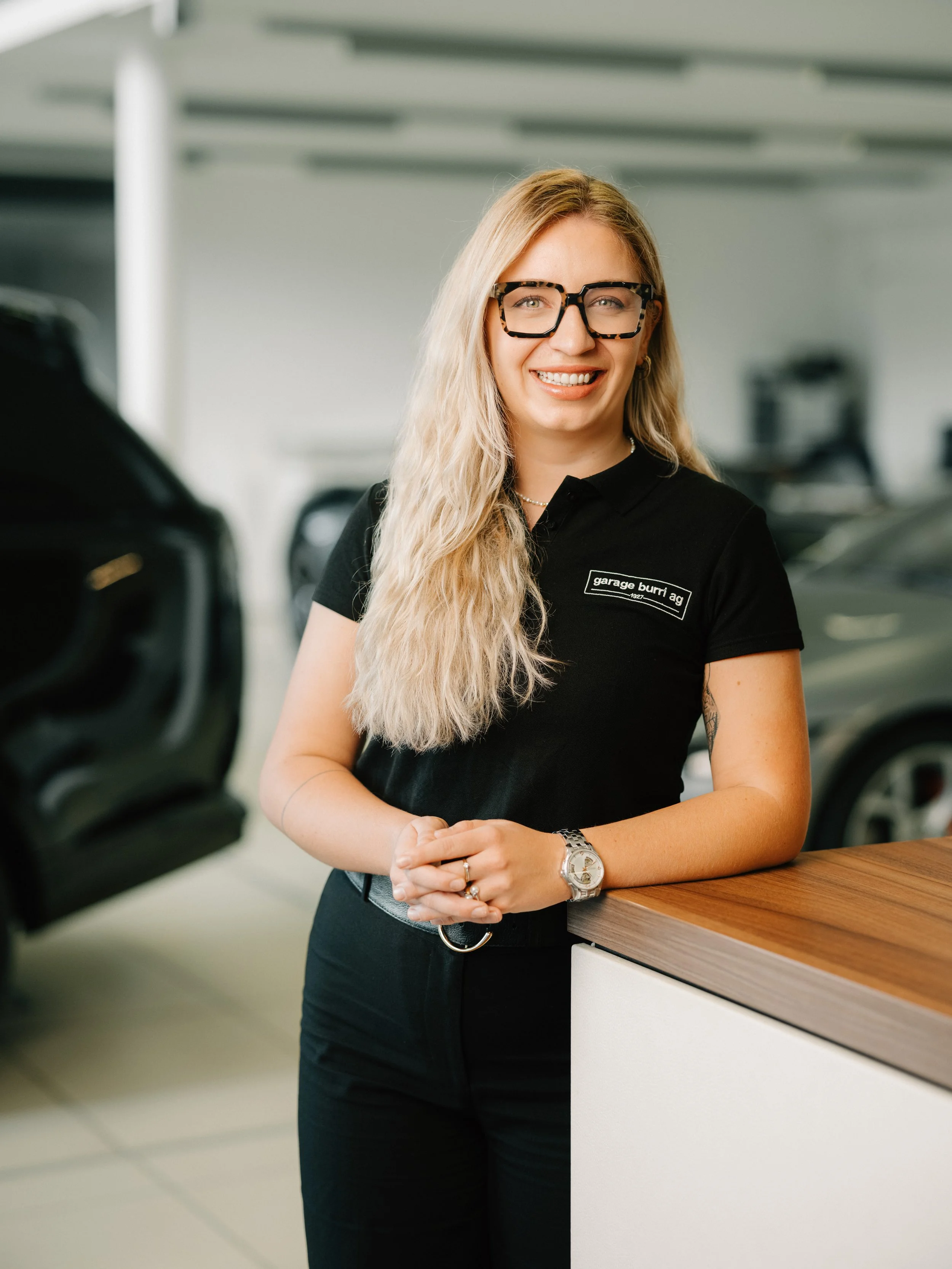 Junge Frau mit Brille, blond, im weißen Polo-Shirt, lächelnd, vor einem modernen, texturierten Hintergrund.