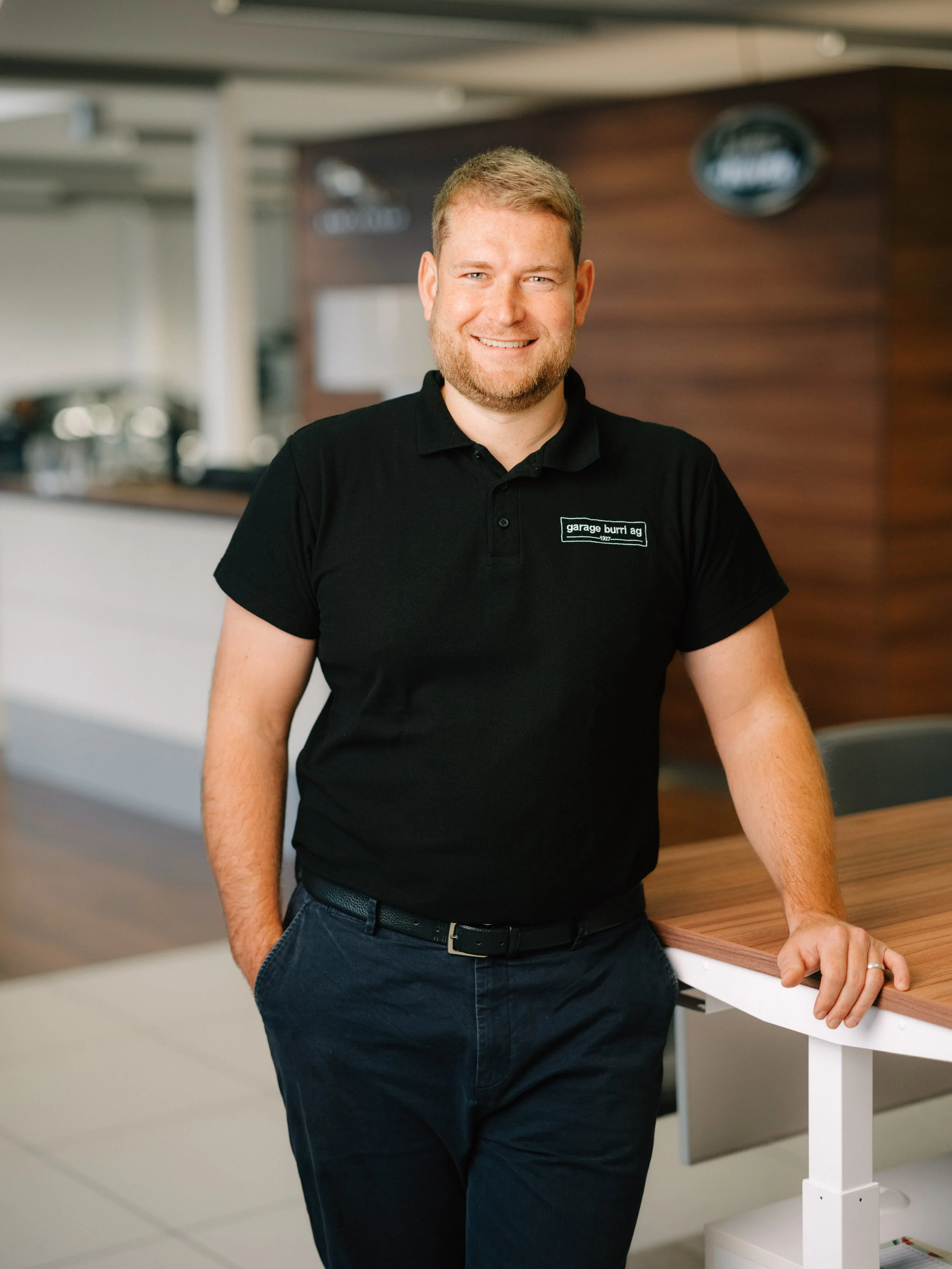 Ein Mann mit kurzen Haaren und Rauschebart trägt ein weißes Poloshirt vor einem hellen, geometrischen Hintergrund.