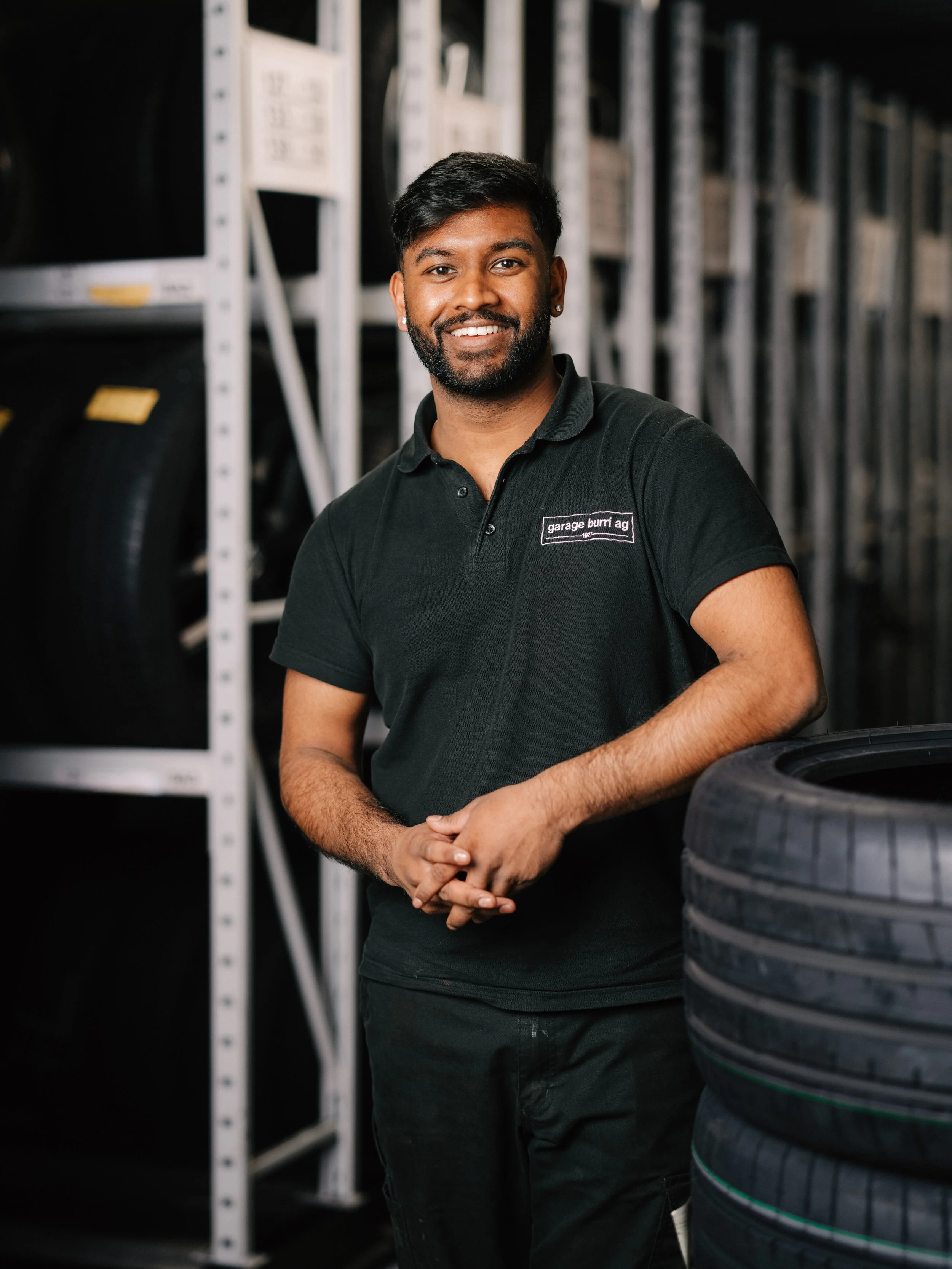 Lächelnder Mann mit dunkler Haut, schwarzem Haar, Bart und weißem Hemd vor einer weißen, modernen Wand.