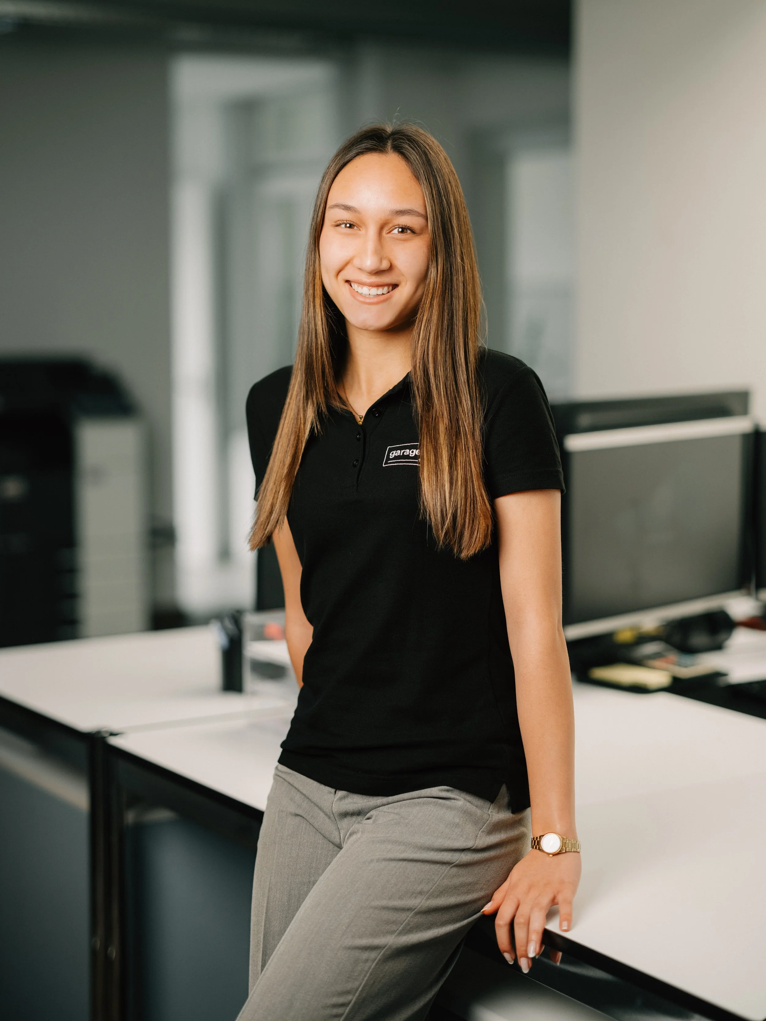 Junge Frau trägt weiße Bluse, lächelt, mit dunklen langen Haaren, Hintergrund mit gleichmäßigen, hellen, geometrischen Mustern.